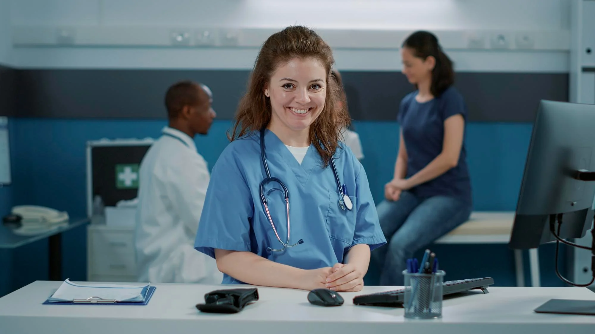 A female healthcare professional, wearing blue scrubs and a stethoscope, smiling at the camera in a hospital or clinic setting, with other medical staff and a patient in the background.