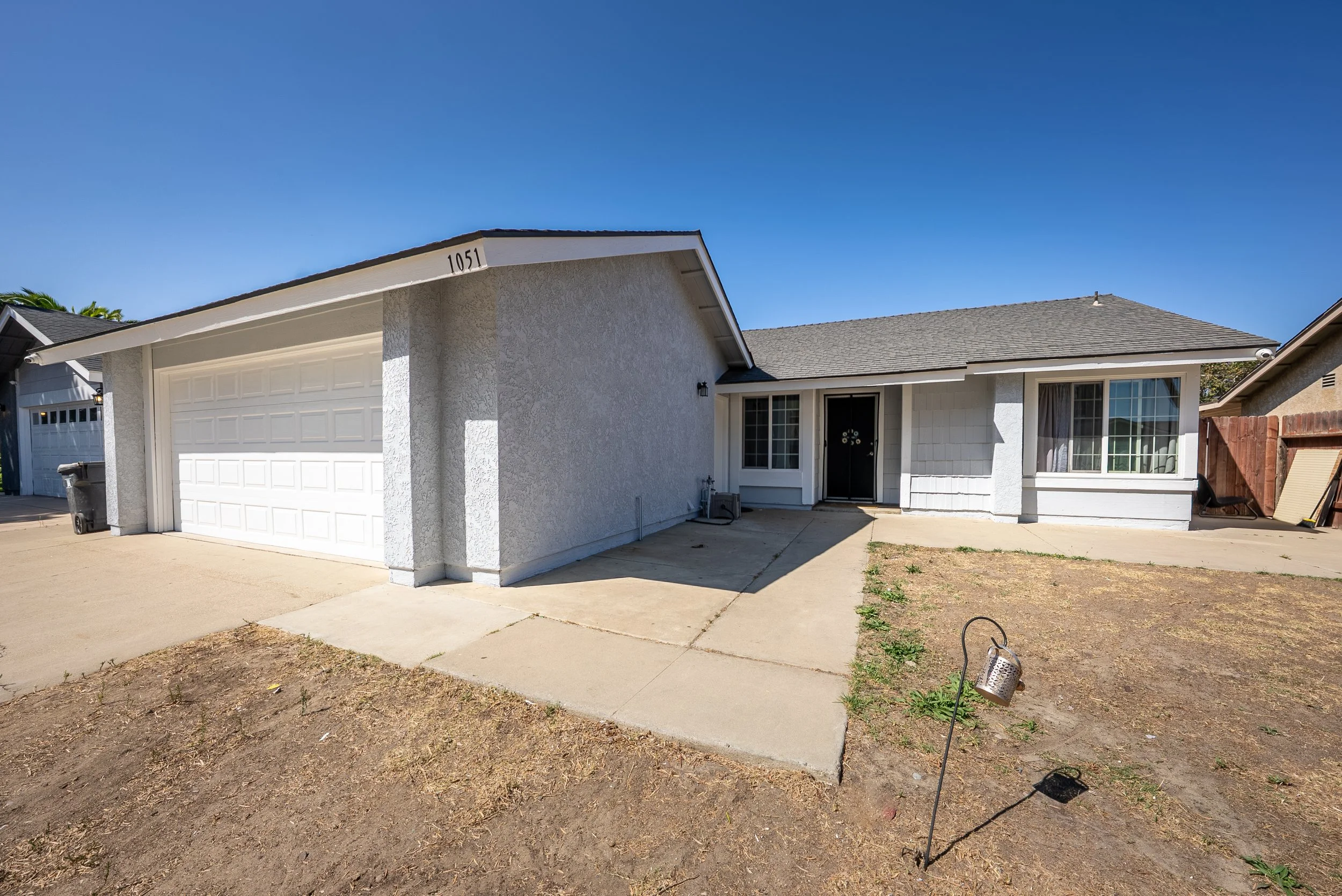 Front view of a white single-story house with an attached garage, a concrete driveway, and a small front yard with bare ground, against a clear blue sky.