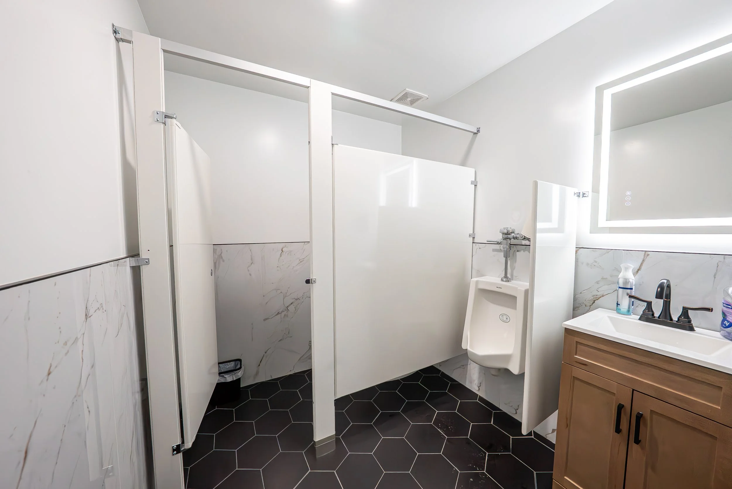 A modern women's public restroom with white stalls, a small sink, and a black hexagonal tile floor.