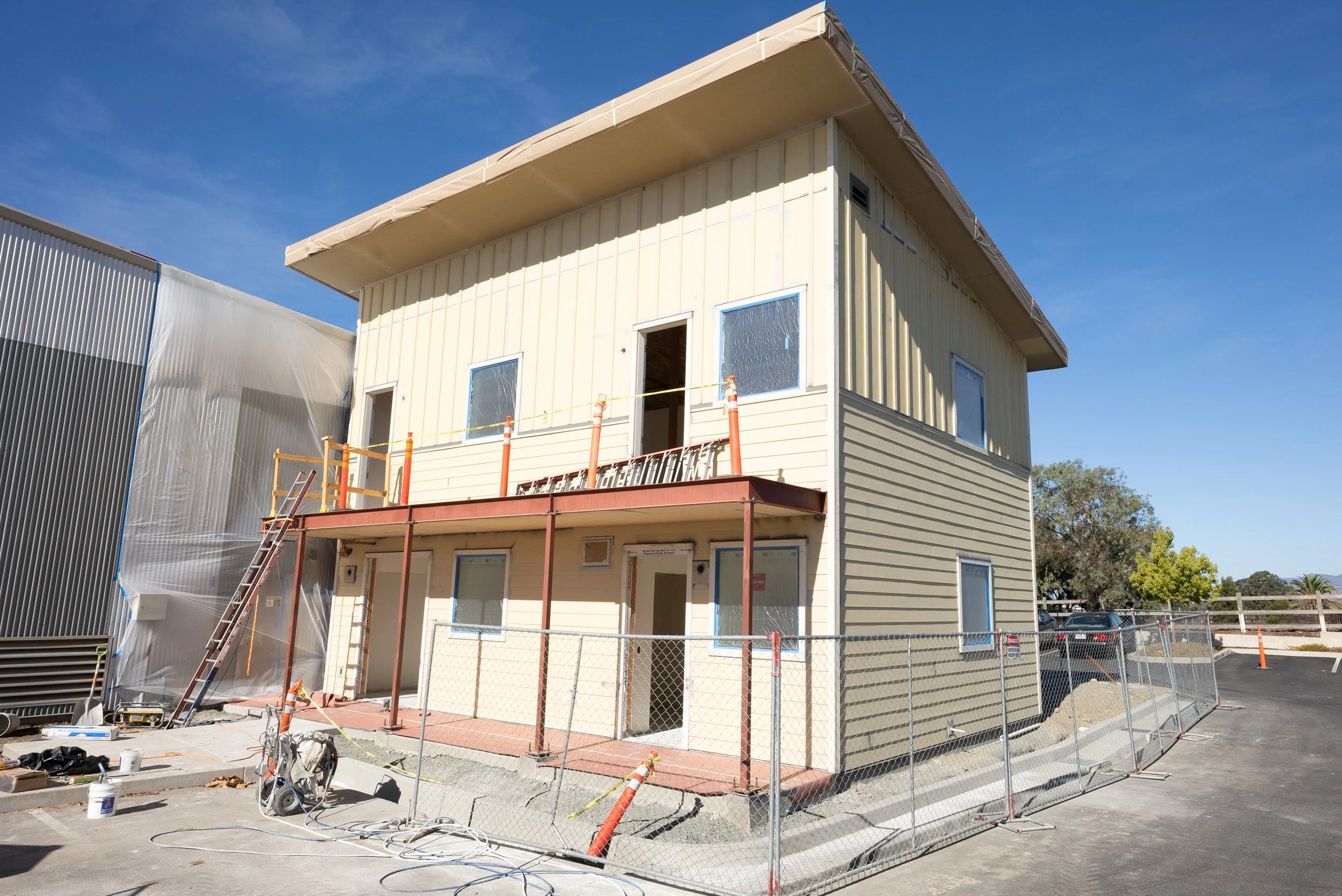 Under construction beige and cream-colored house with scaffolding and construction cones in front, surrounded by a chain-link fence, under a clear blue sky.