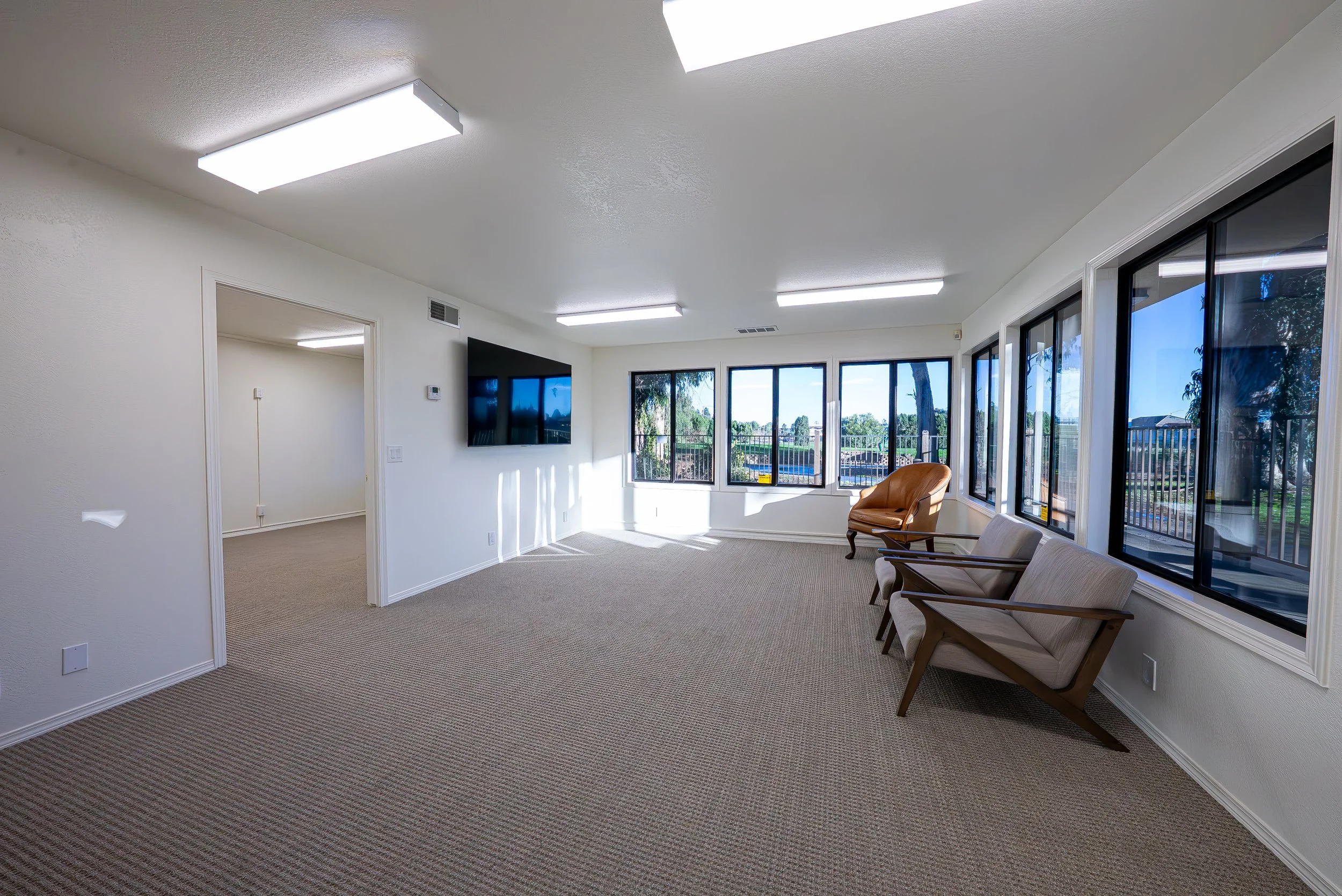 Spacious living room with large windows, beige carpet, white walls, a wall-mounted flat-screen TV, and a few chairs, overlooking a backyard with trees and a swimming pool.