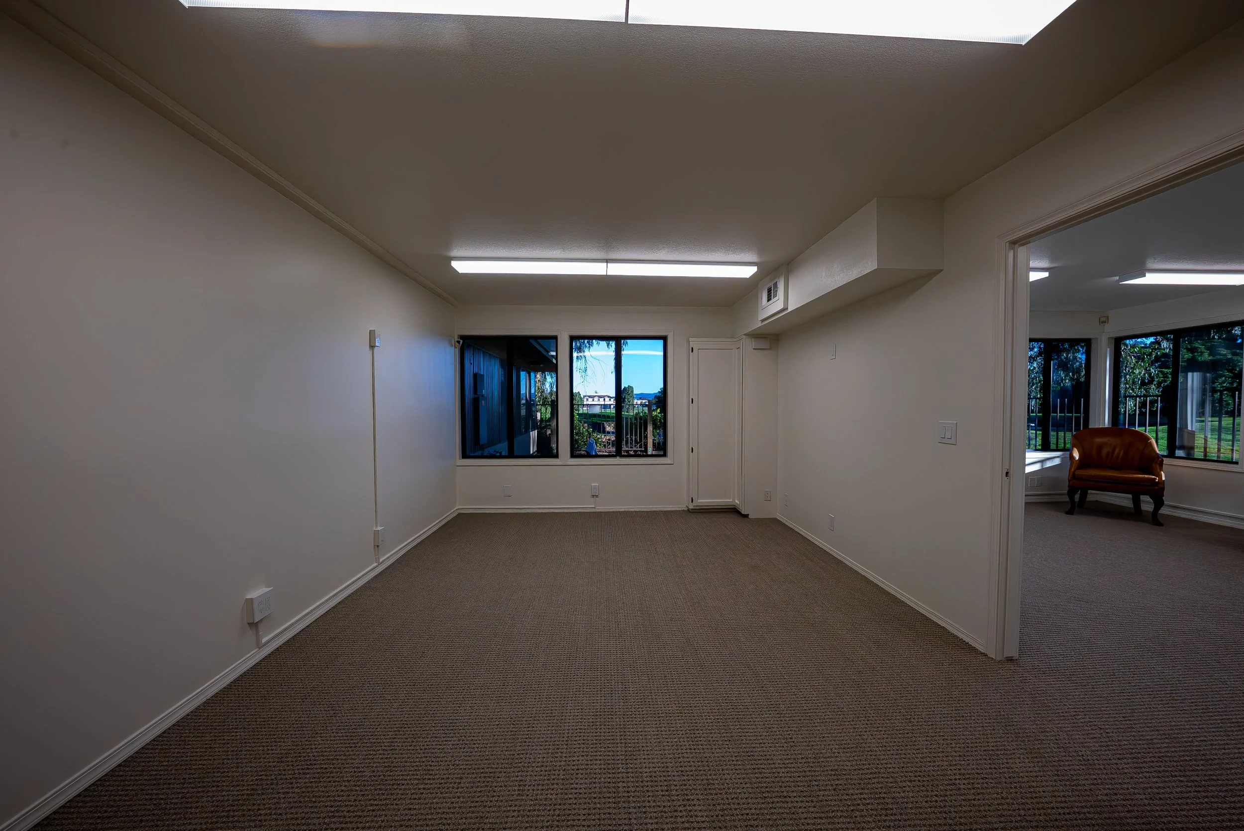 Empty living room with beige carpet, white walls, and a large window overlooking a balcony and greenery outside. Fluorescent ceiling lights, doorway to another room with additional large windows, and an orange armchair are visible.