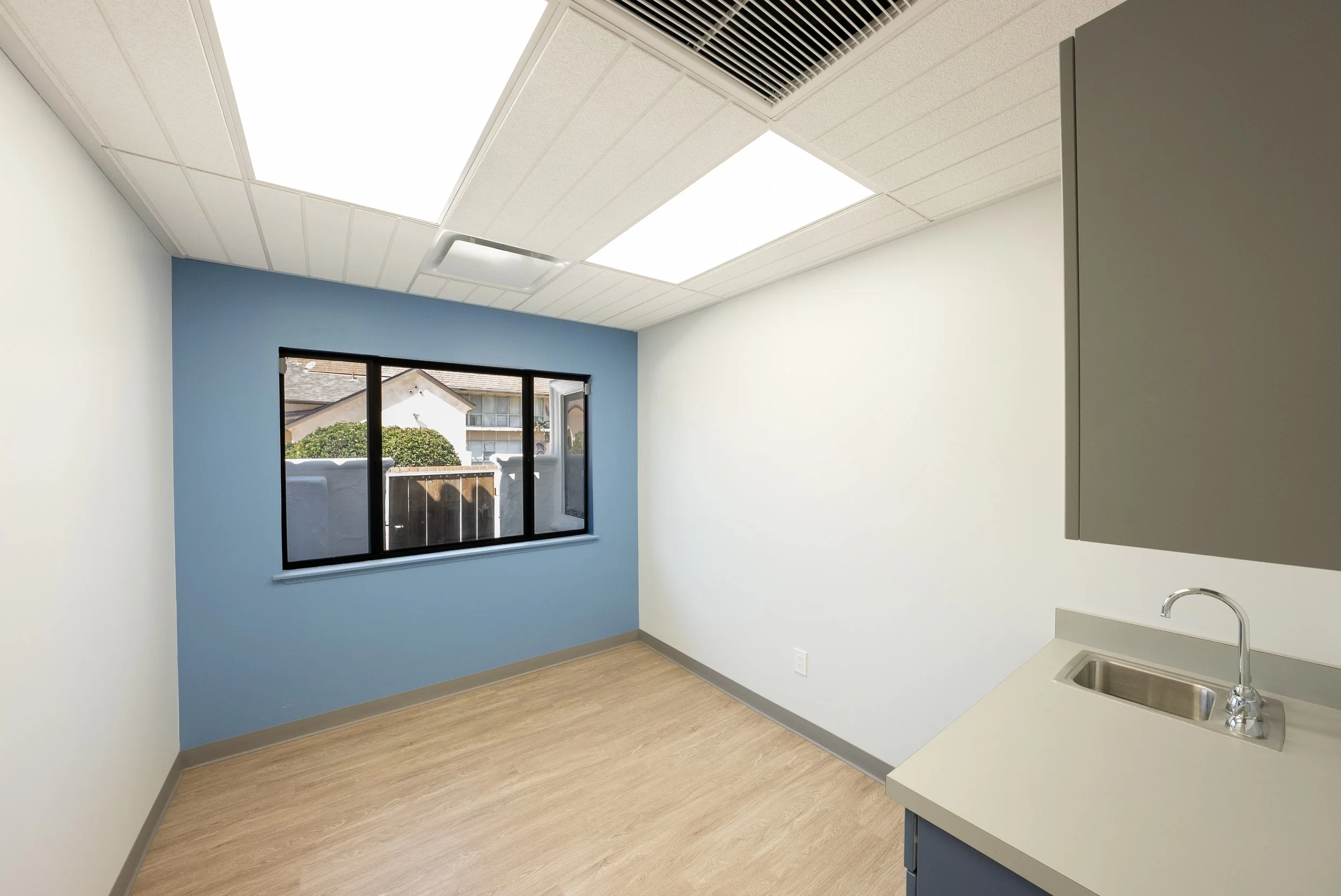 Empty room with white walls and a blue accent wall, wood flooring, a large window with a view of neighboring houses, ceiling lights, and a small section of a kitchen counter with a sink.