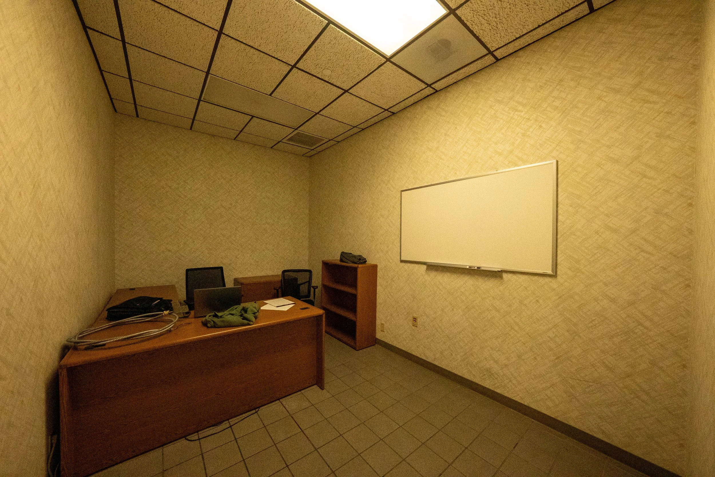 An empty office with beige patterned walls, a ceiling with square tiles and fluorescent lighting, a wooden desk with office supplies, two chairs, a small bookshelf, a blank whiteboard, and tiled flooring.