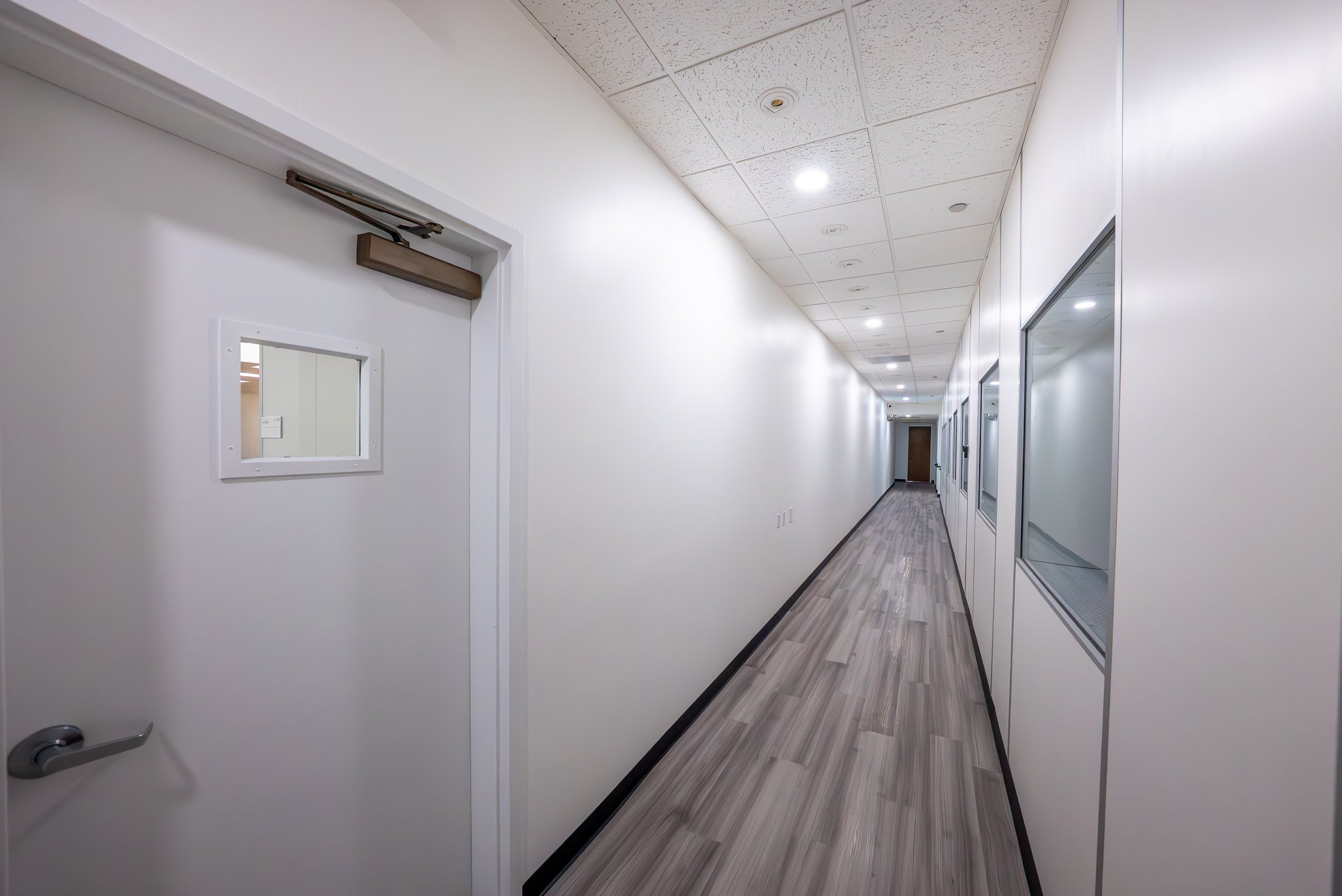 Long hallway with white walls, ceiling panels, and wood-look flooring, featuring windows on the right side and a door with a small mirror on the left side.