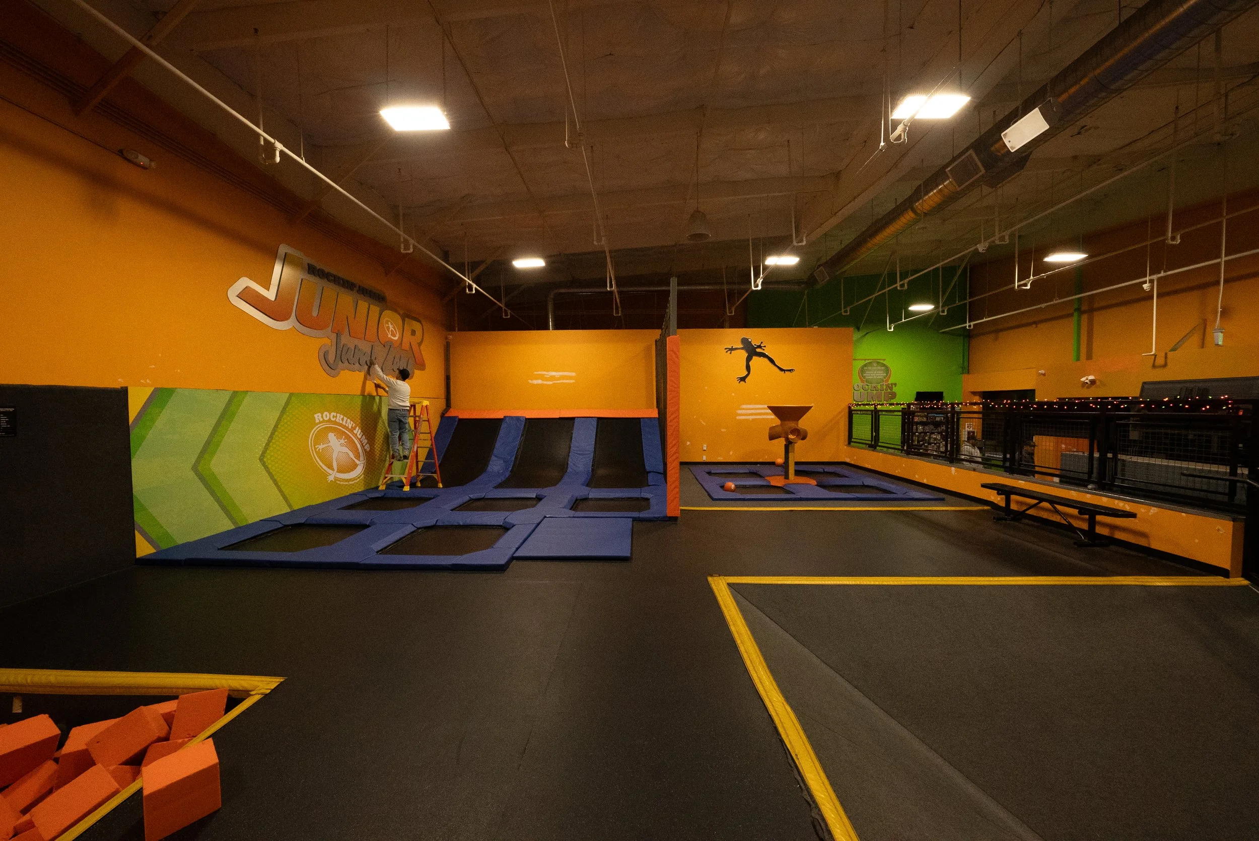 An indoor trampoline park with a person standing on a ladder near a wall, which has the words "DUNK" and "JUMP" on it. The facility includes multiple trampoline sections with padded surfaces and a large wall with a silhouette of a person jumping. The