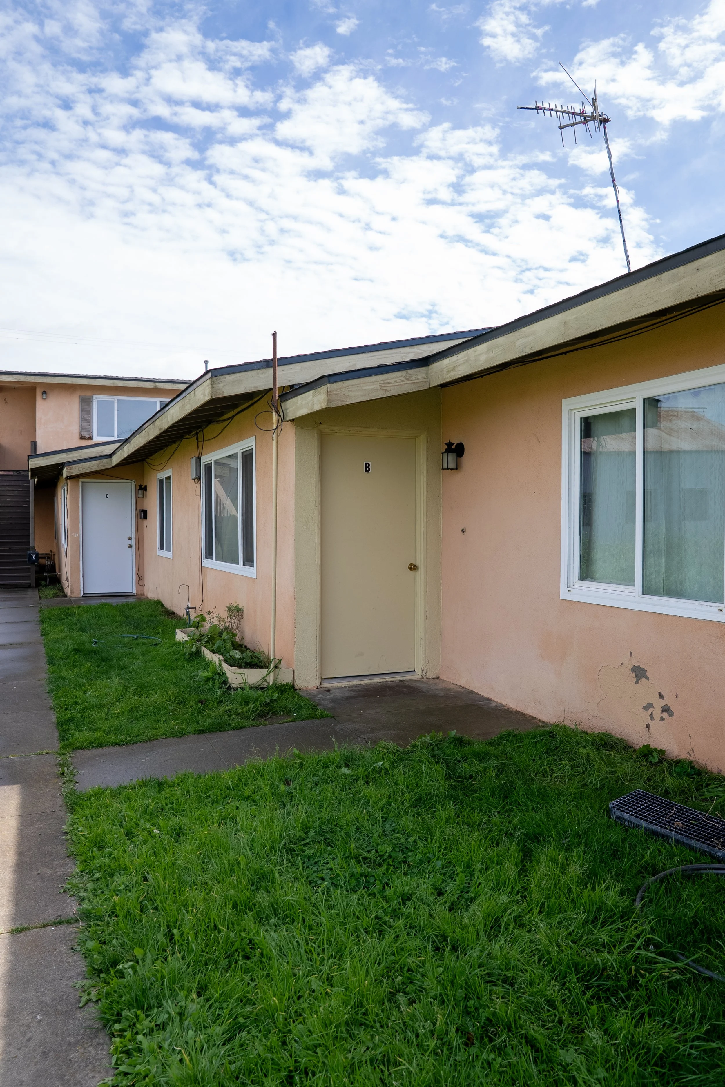 Front view of a peach-colored apartment building with white doors and windows, a small lawn, and an overgrown sidewalk. The sky is partly cloudy with a television antenna visible on the roof.