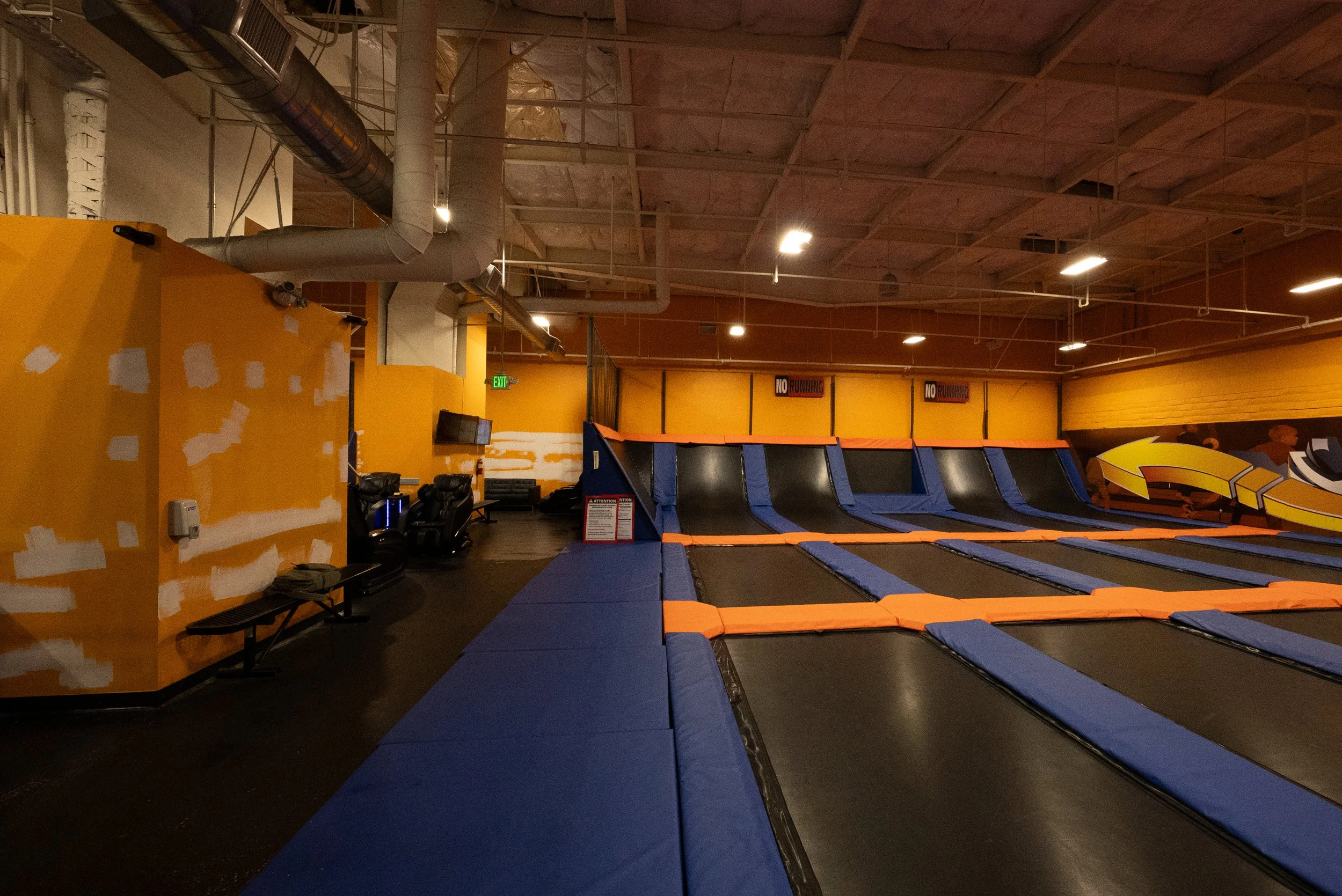 Indoor trampoline park with multiple blue and orange trampolines arranged in a row, yellow and black walls, industrial ceiling with pipes, and signs that say 'No Running'