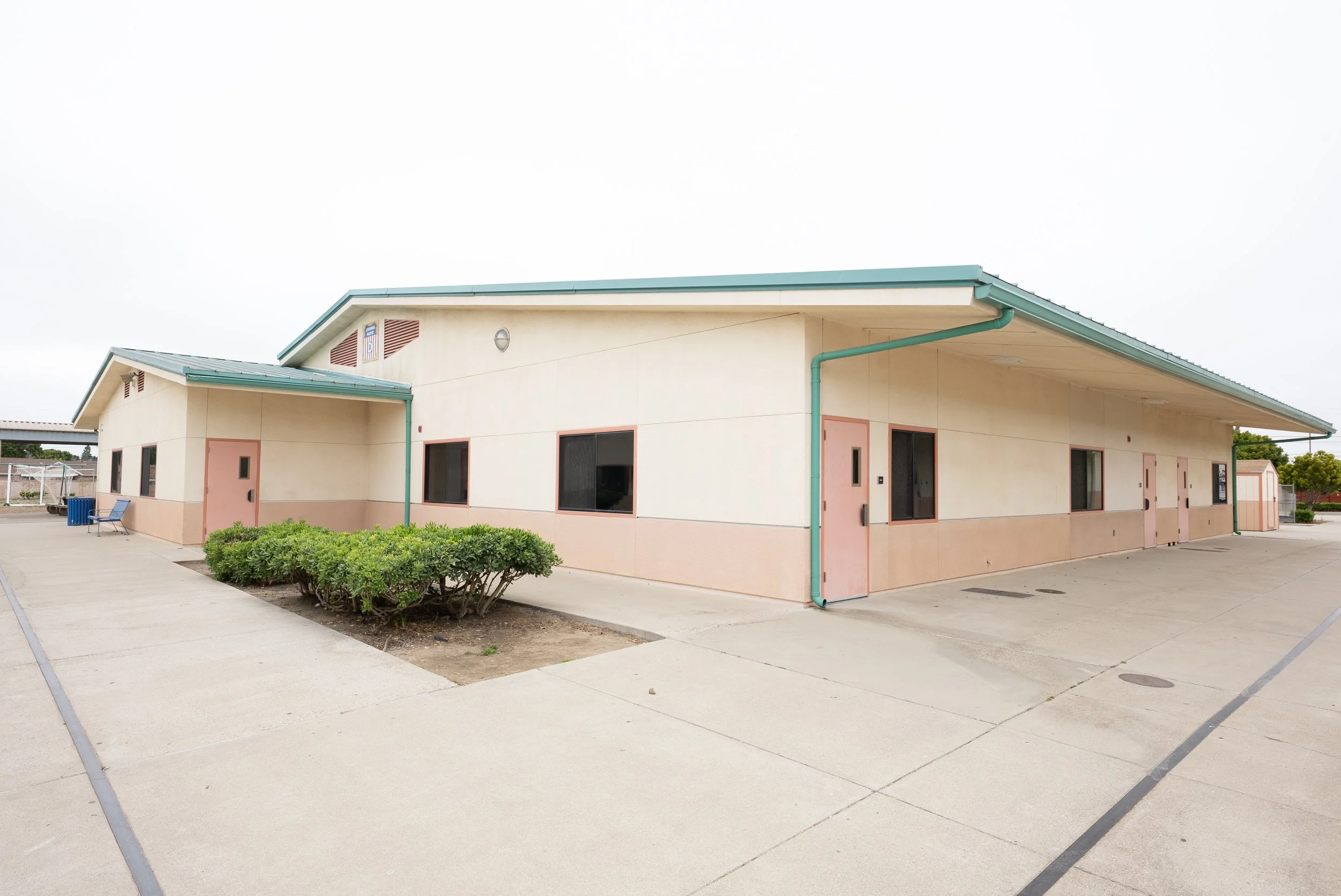 A single-story building with a light-colored exterior, teal roof, and matching teal rain gutters. It has several windows and doors, with some shrubbery in front. There are empty benches and a paved area surrounding the building, under an overcast sky