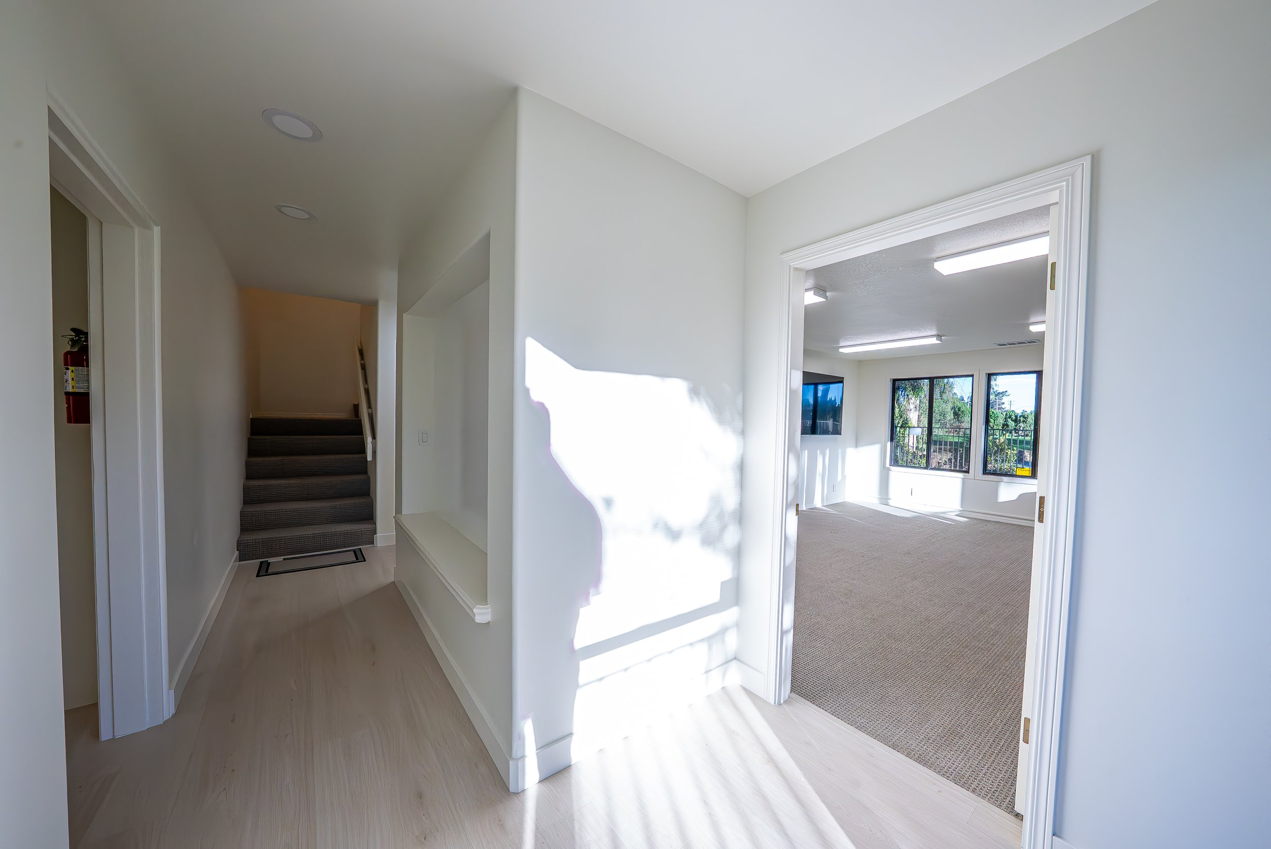 Interior view of a bright hallway with a staircase on the left, leading upward, and a doorway on the right opening to a room with carpeted floor and large windows showing outdoor scenery.