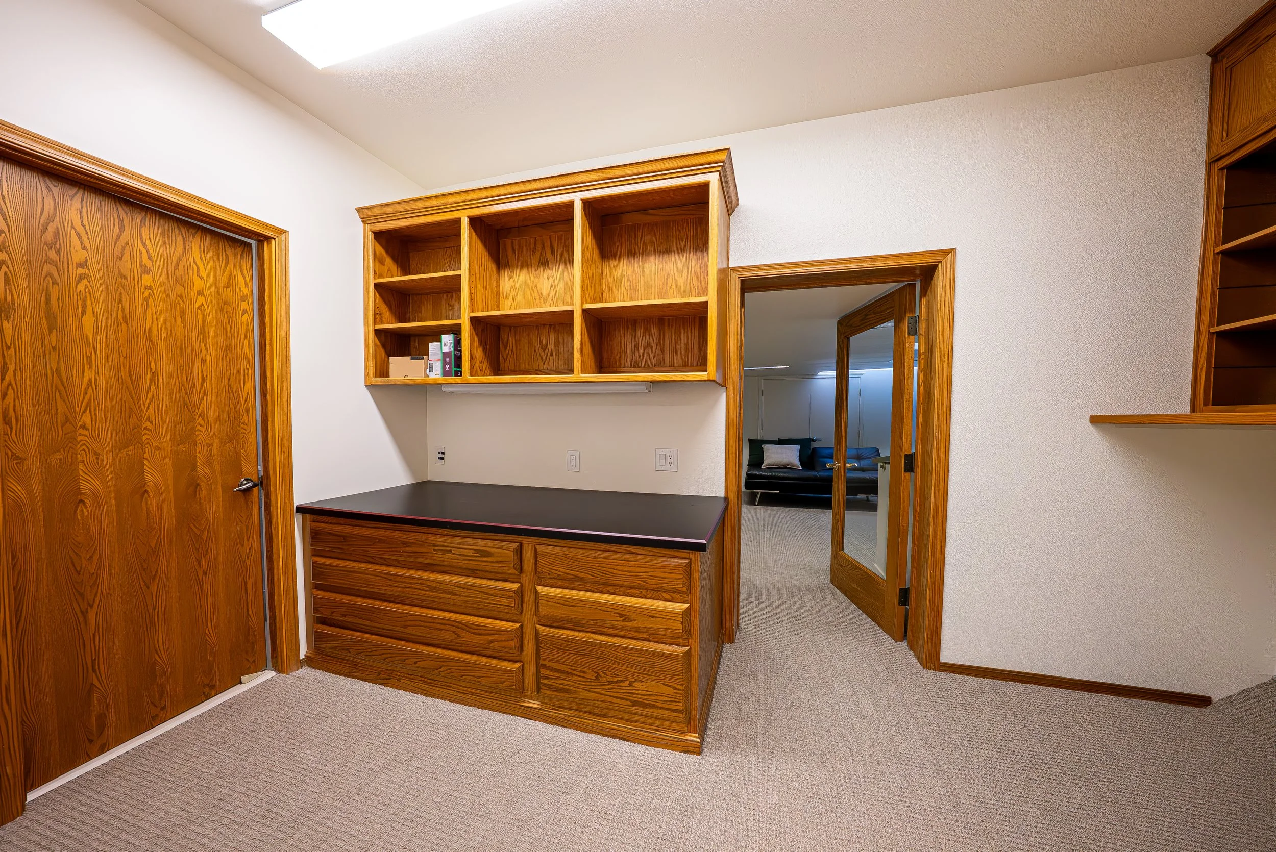 A room with wooden cabinets, a door, a doorway leading to a living room with a couch and pillows, and ceiling skylight.