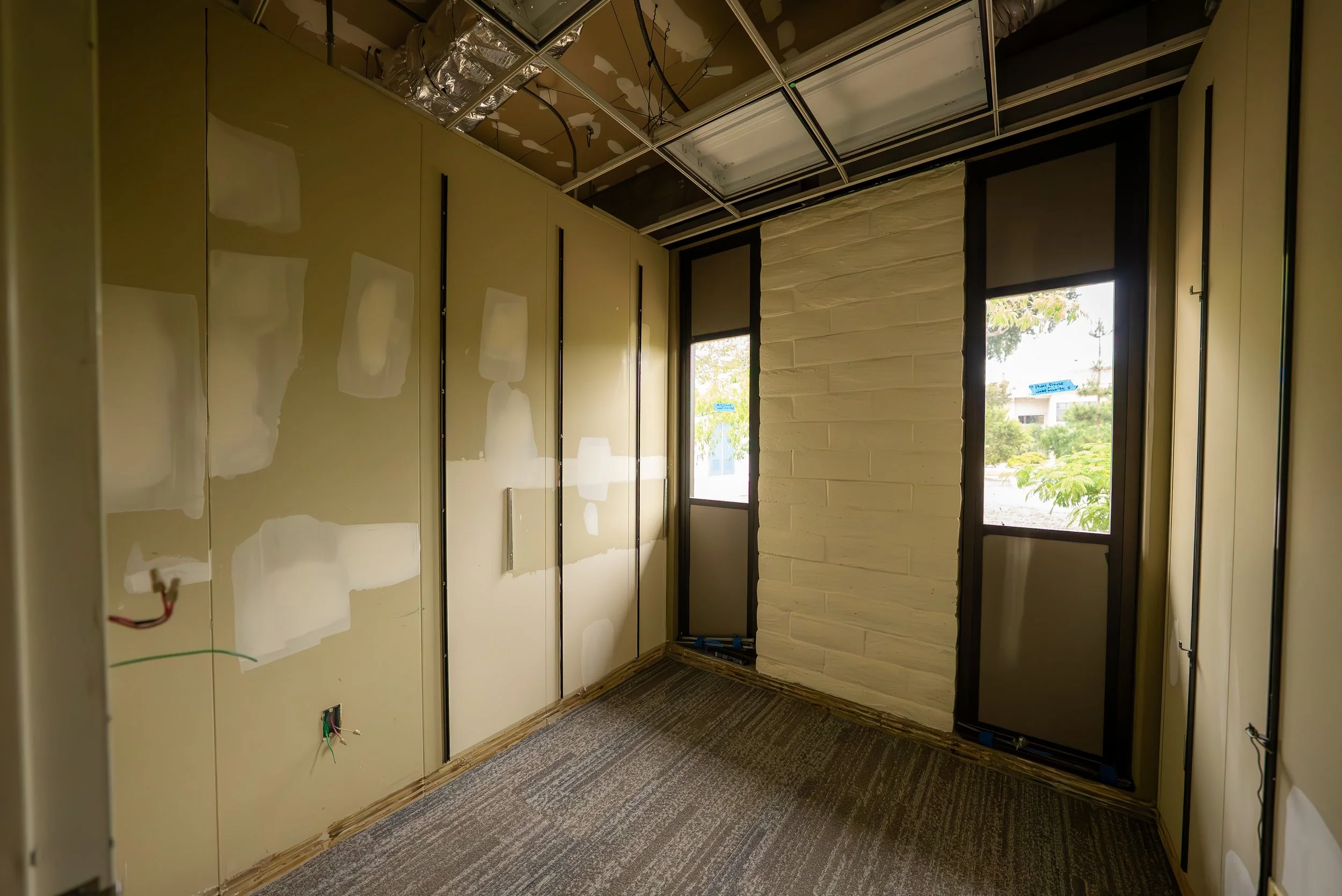 Interior of an unfinished room with drywall panels, electrical wiring, and a window showing trees outside.