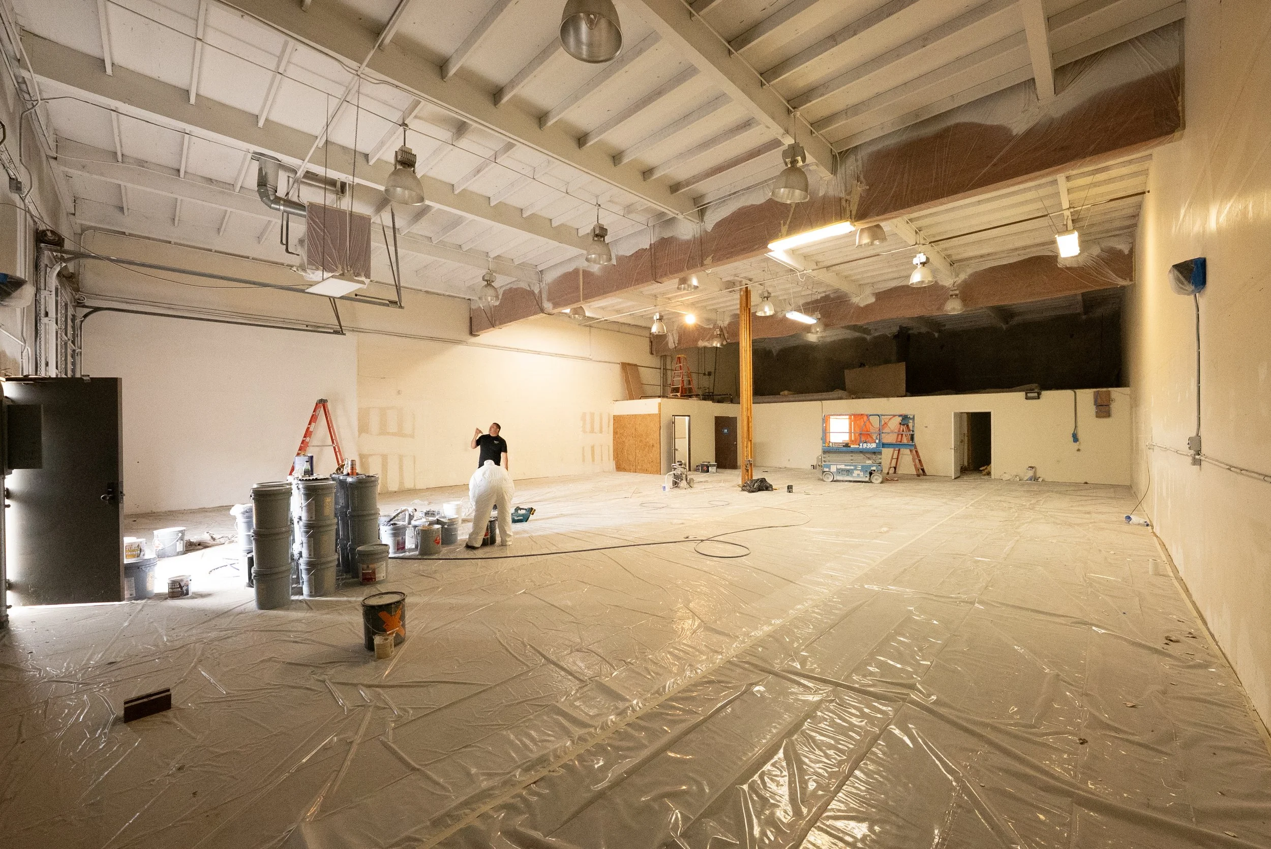 Interior of a large room under renovation, with construction materials, ladders, and workers present. The ceiling is exposed with insulation and lighting fixtures installed. The floor is covered in plastic sheeting.