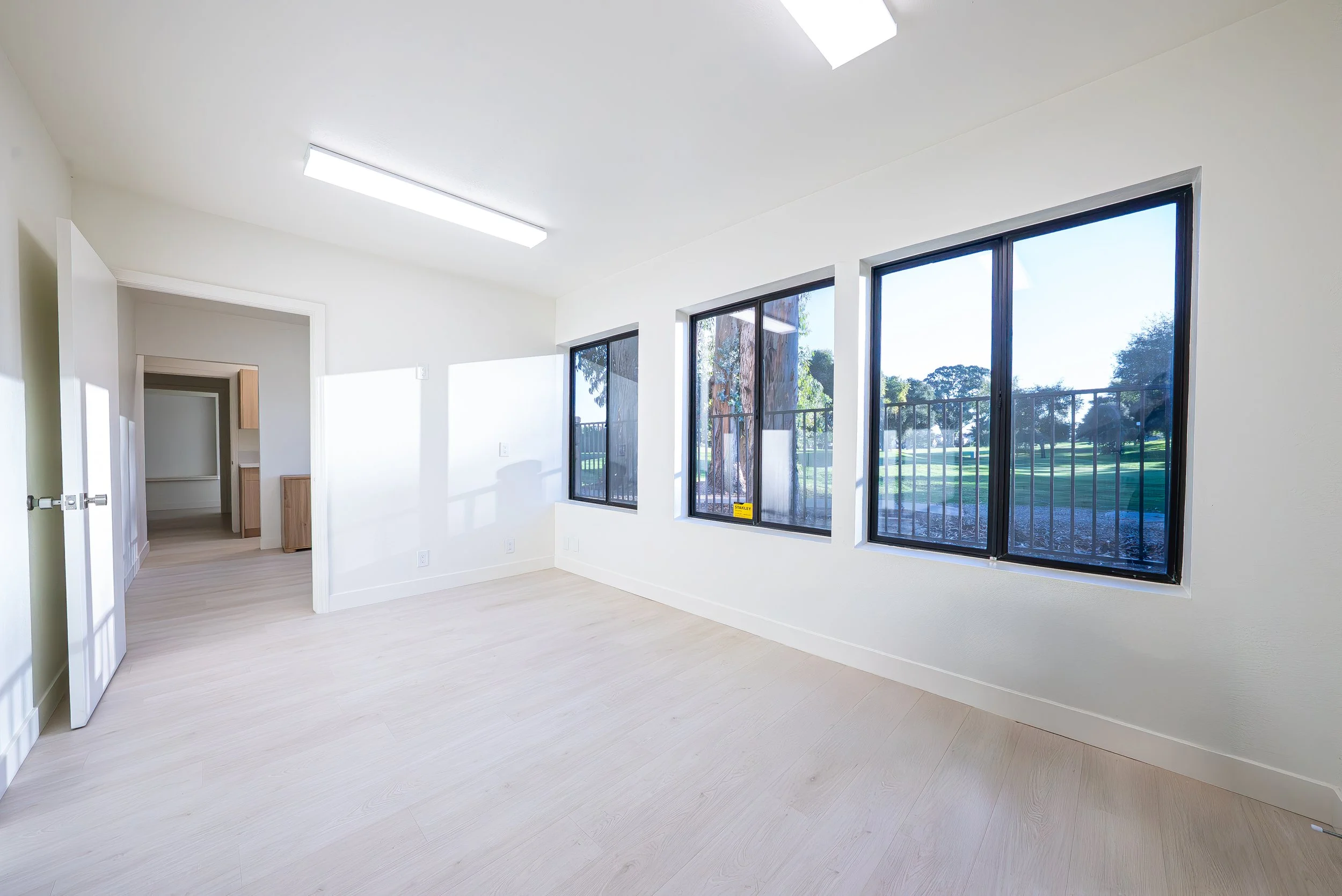 Empty room with light-colored wooden floors, large windows showing trees outside, and white walls.