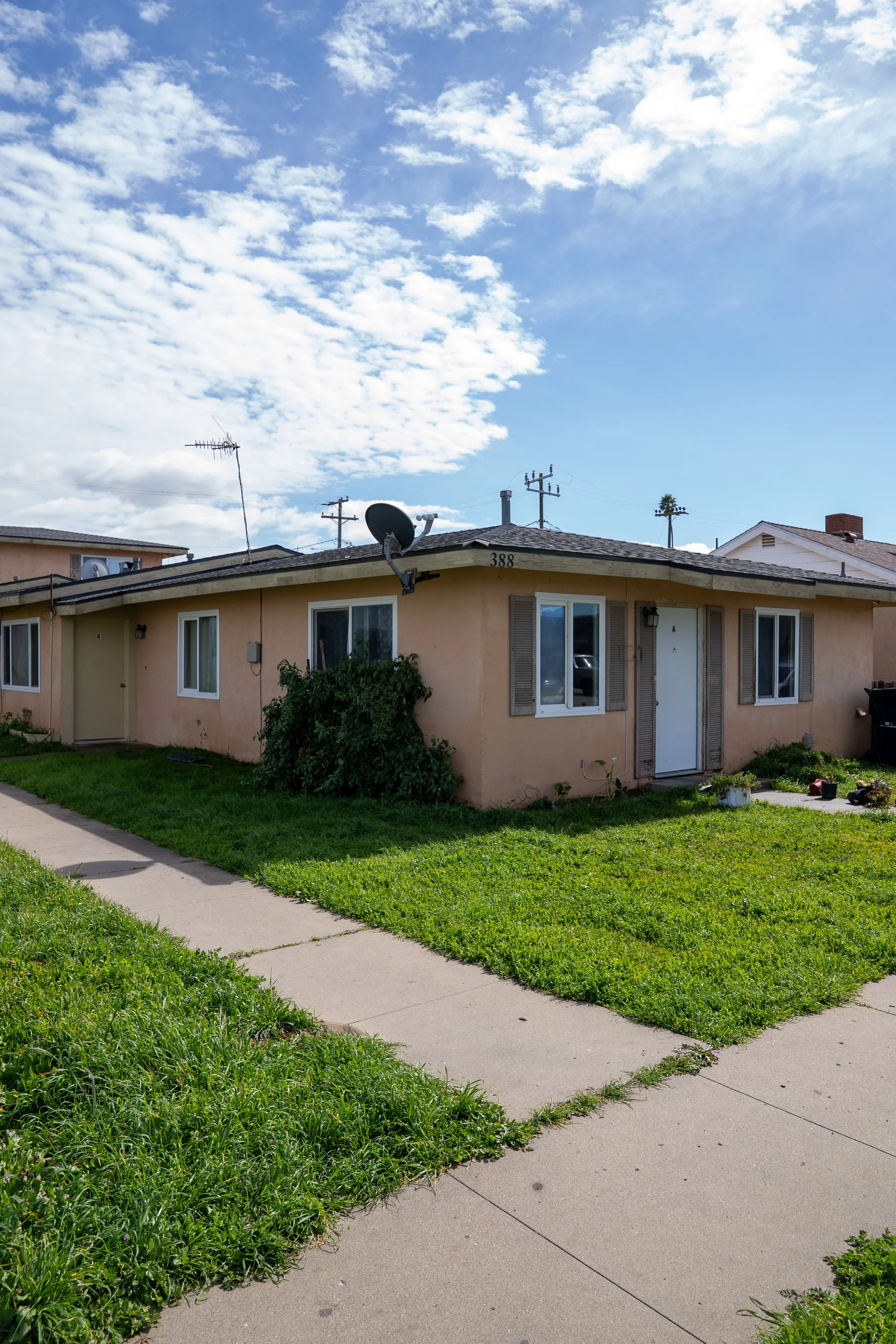 A single-story beige house with a flat roof, satellite dish, and windows, surrounded by a grassy lawn and a concrete walkway under a partly cloudy sky.