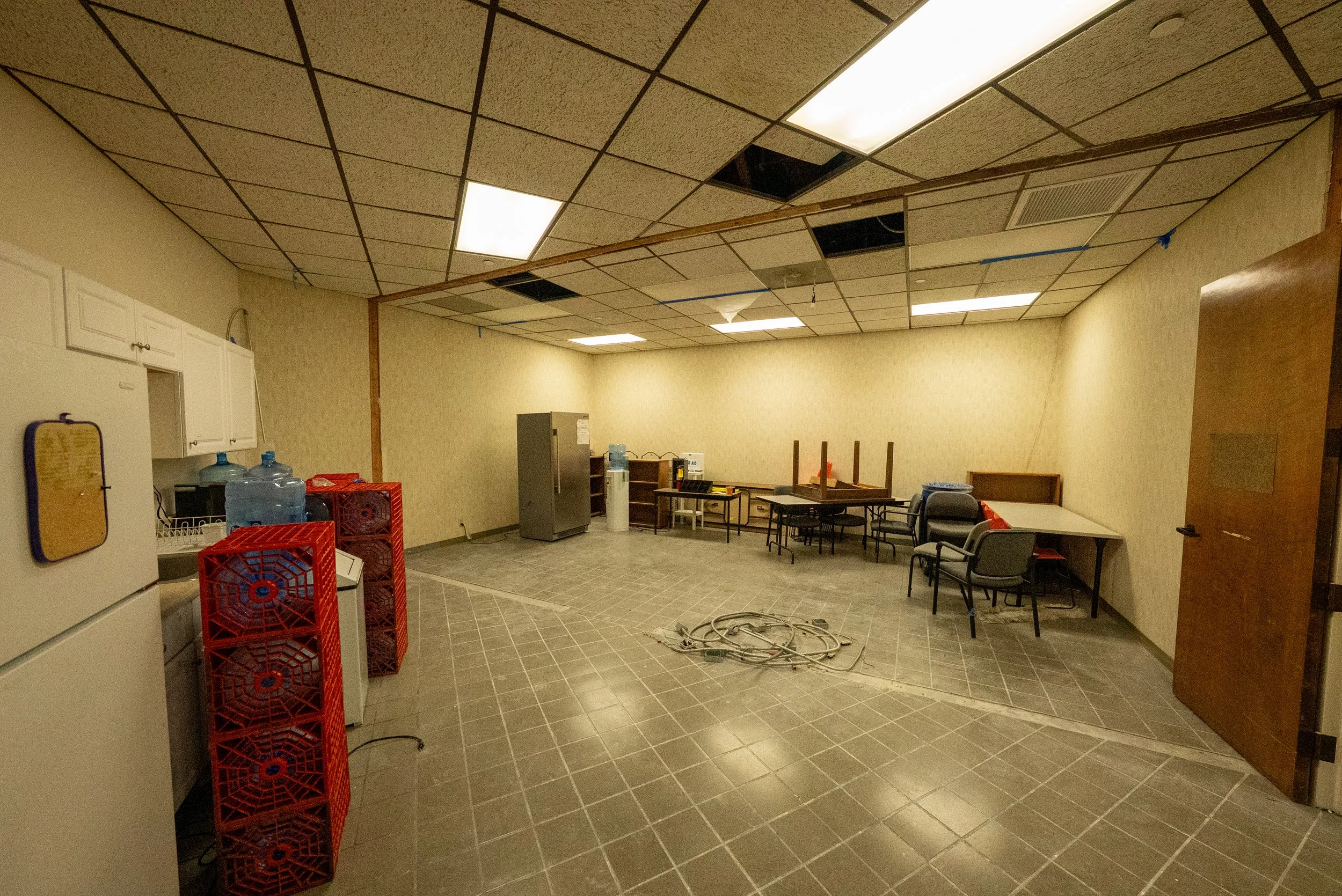 An empty office break room with some ceiling tiles removed and exposed black openings, a water cooler, red crates, tables, chairs, and some wires on the floor.