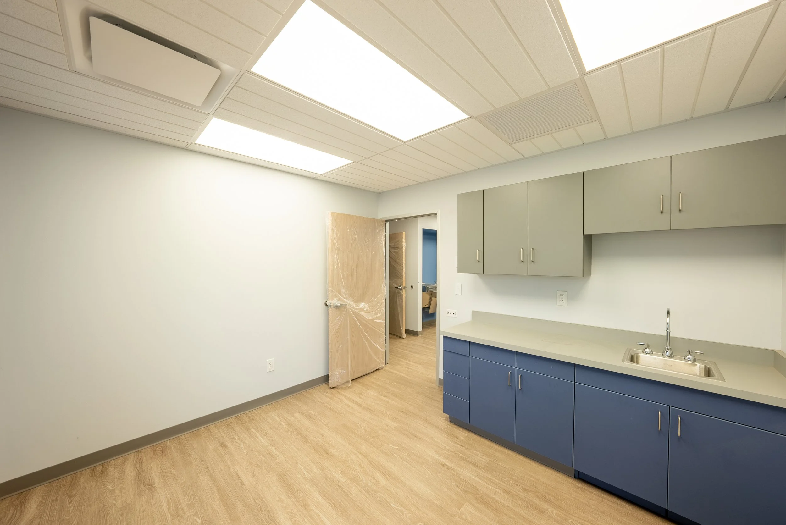 Empty room with a kitchen area, blue and beige cabinets, stainless steel sink, wood flooring, and ceiling lights.