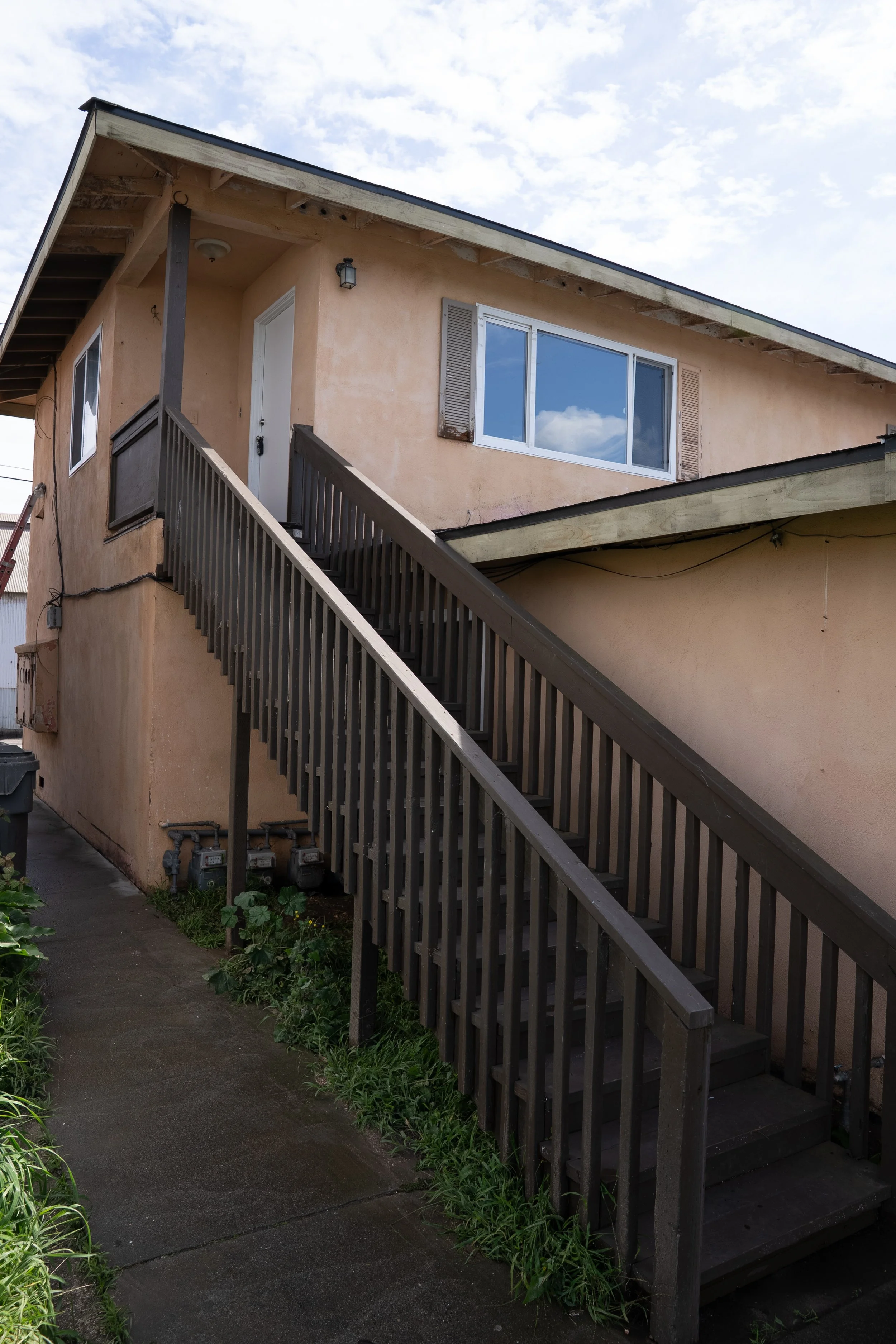 The image shows the exterior of a two-story residential building with beige stucco walls, a dark roof, and an outdoor staircase with a wooden railing leading to the second floor. There is a large window on the second floor reflecting the sky with clo