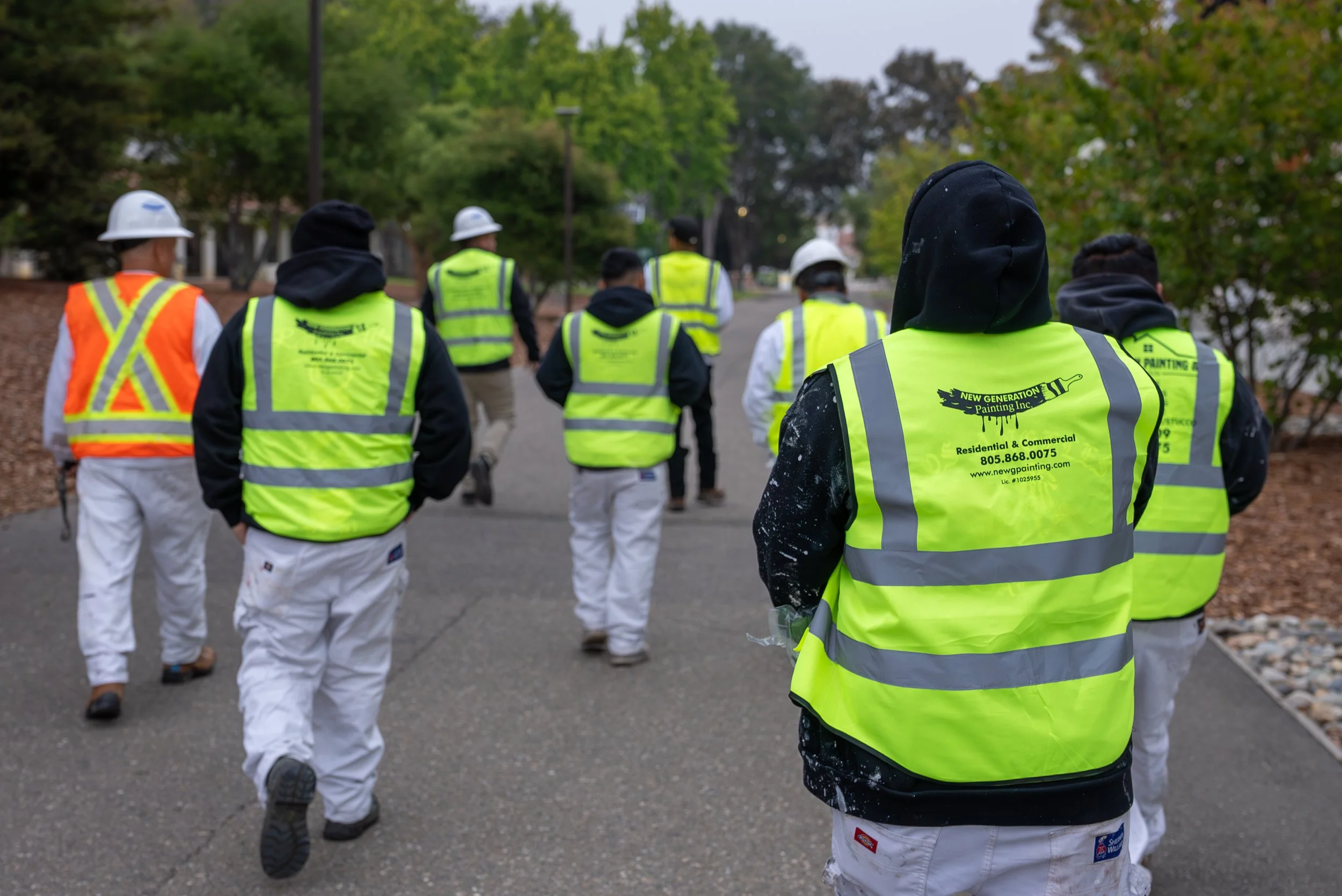Group of construction workers walking along a street in a park-like area, wearing high-visibility vests and some with helmets, with trees and power lines in the background.