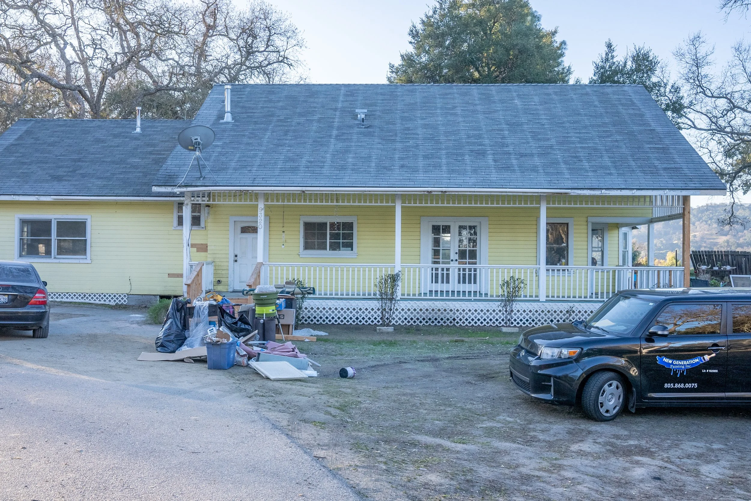 A yellow house with a front porch, missing some steps, and a gray roof. There are several cars parked in the driveway and a pile of trash and debris near the porch. The background features large trees and a clear sky.