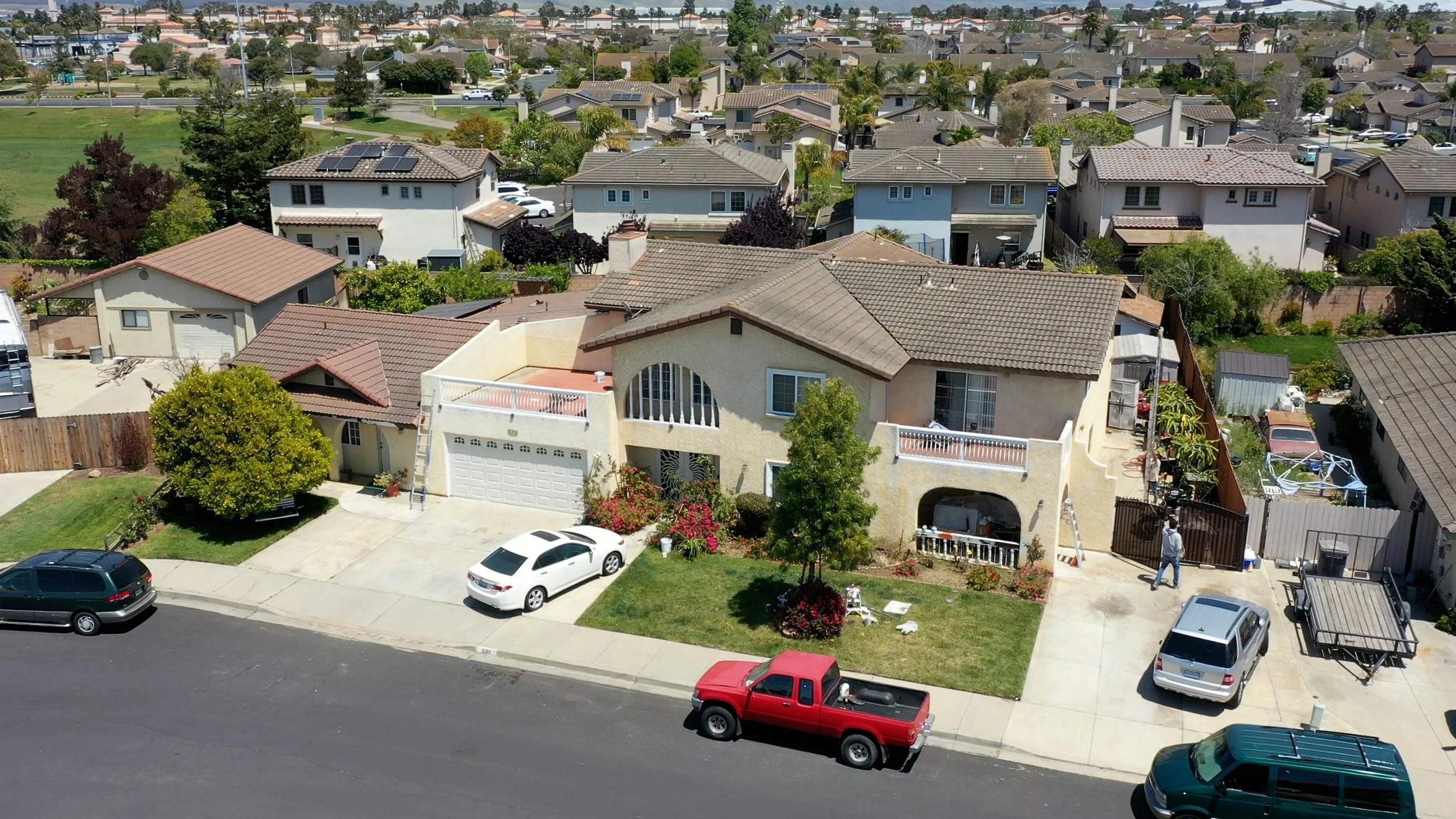 Aerial view of a residential neighborhood with multiple houses, cars parked along the street, green lawns, trees, and a park in the background.
