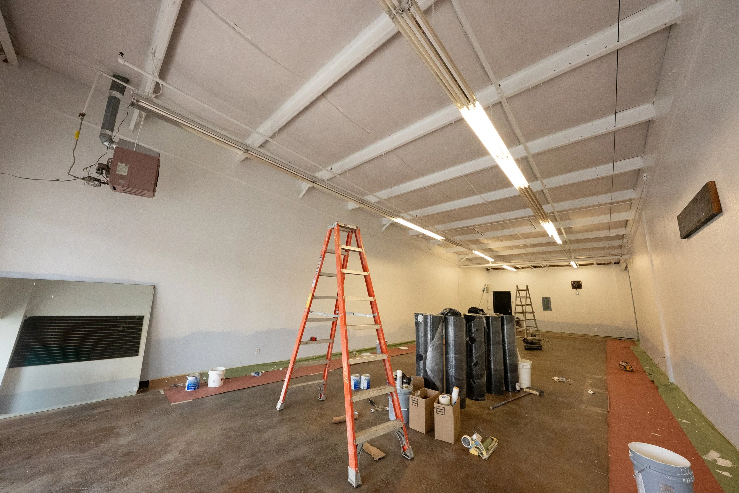 Interior of a room under renovation with exposed ceiling beams and fluorescent lights. Construction materials, ladders, and paint supplies are scattered across the floor.