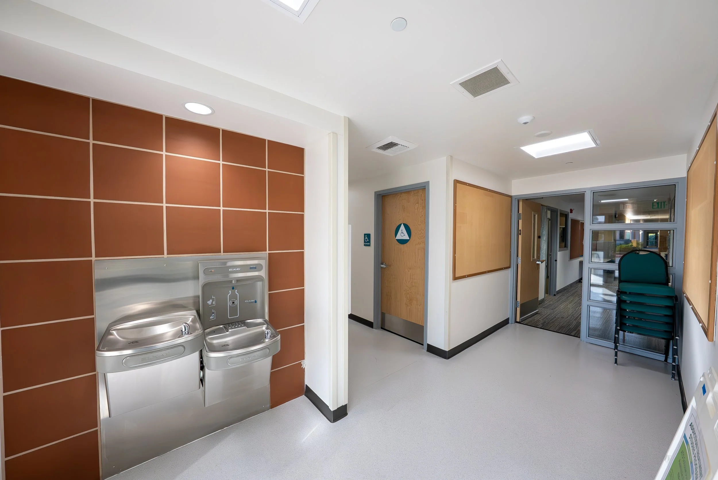 Interior of a hospital or clinic with a drinking fountain, brown tiled wall, wooden and glass doors, blue chairs, and a bulletin board on the wall.