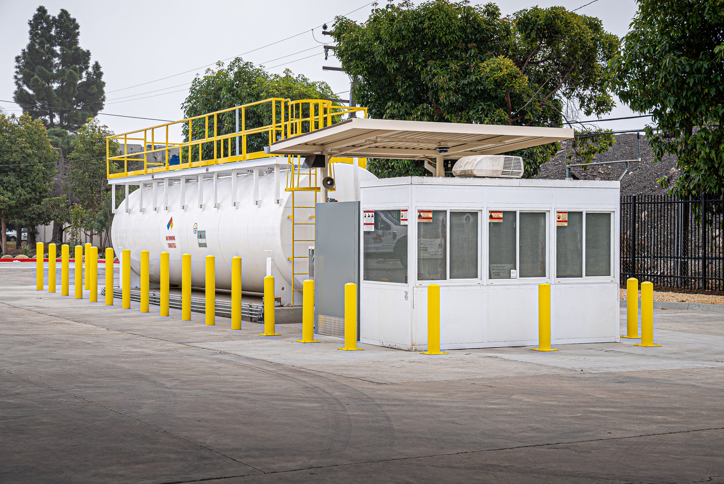 A propane tank with a small security booth in front, surrounded by yellow safety posts. Trees and power lines in the background.