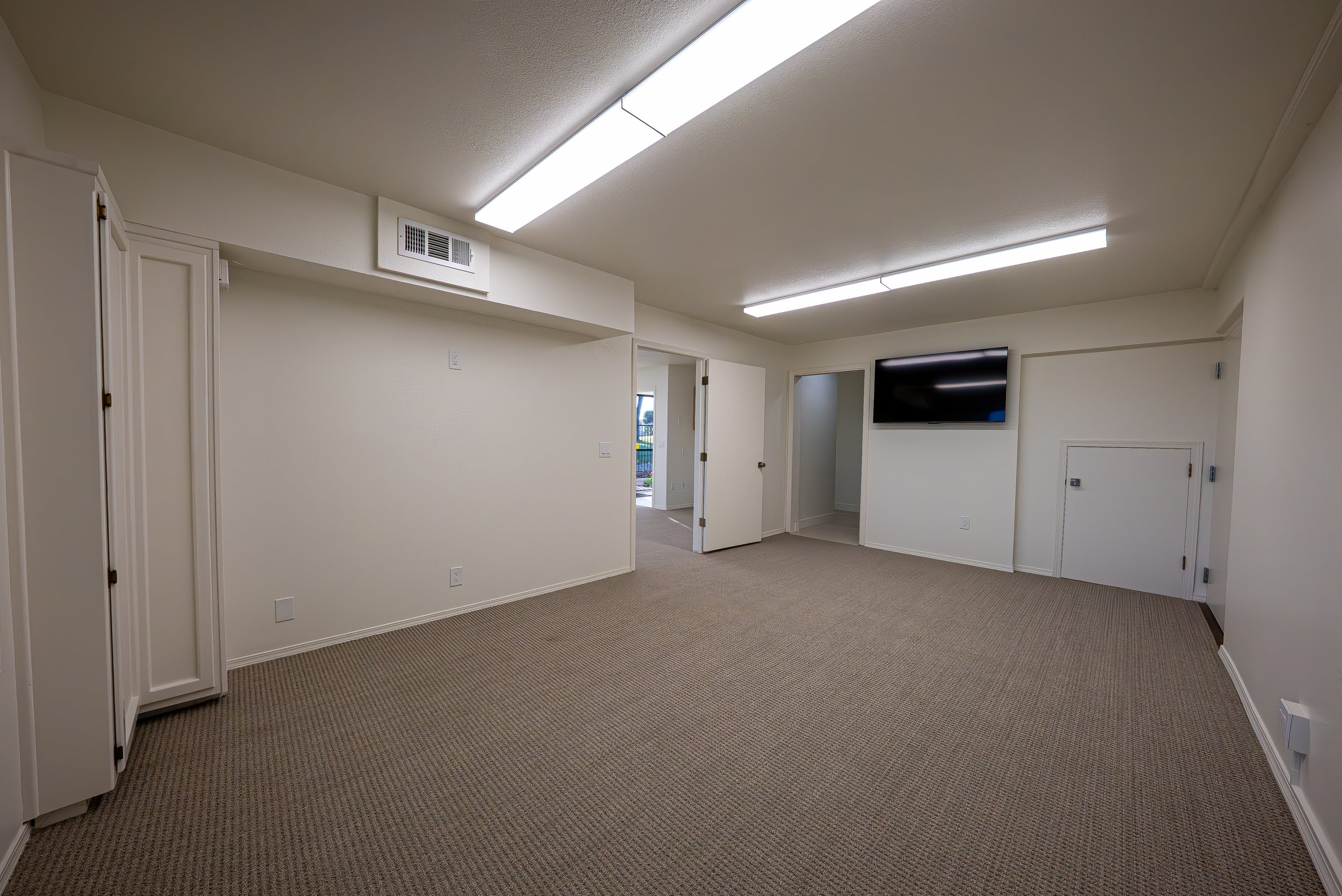 Interior of a room with beige wall-to-wall carpet, white walls, fluorescent ceiling lights, and a mounted flat-screen TV. There are two open doors leading to other rooms and a window showing an outdoor view.