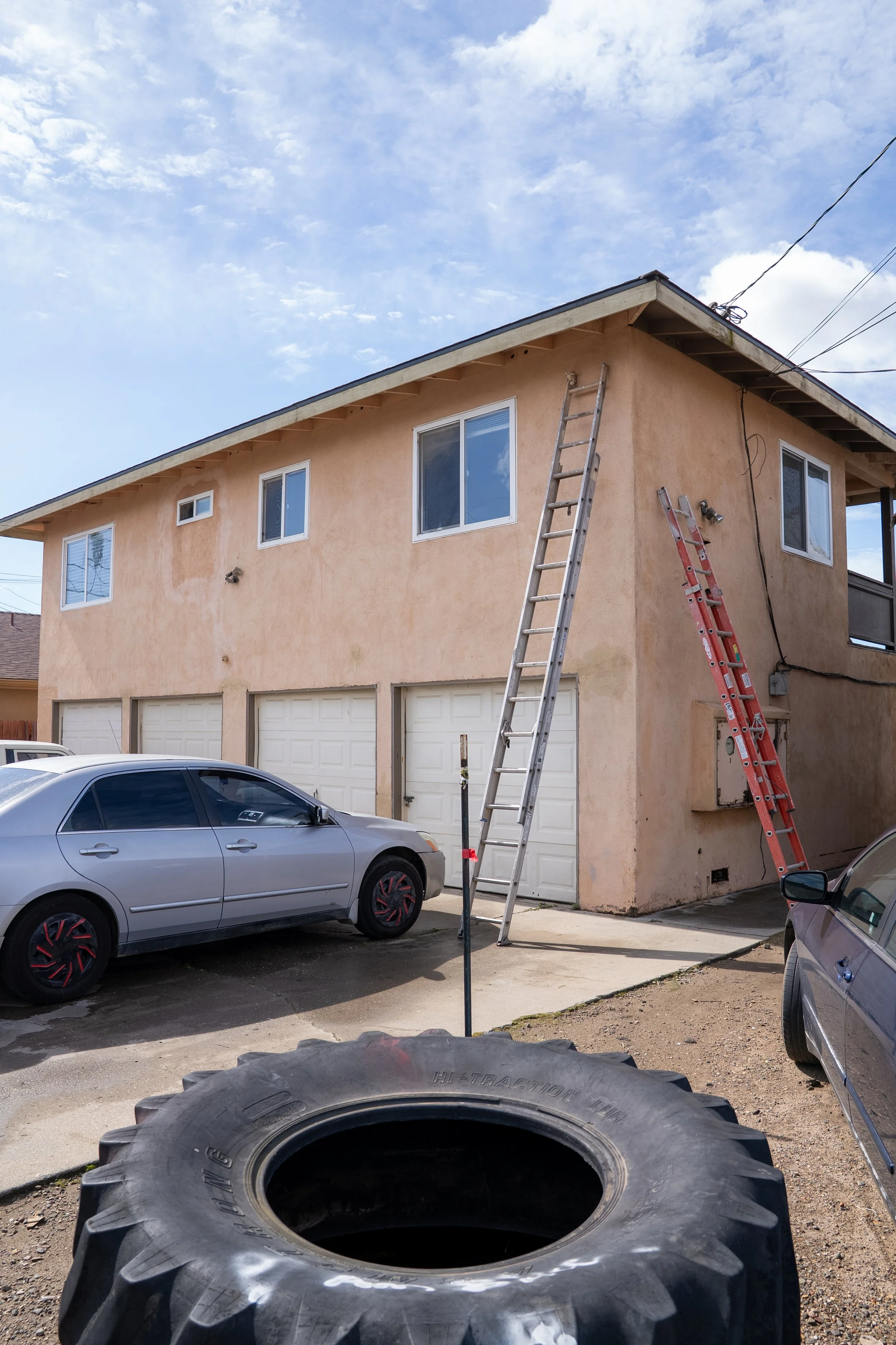 Two ladders leaning against a two-story beige house with a garage. Several cars parked in the driveway. Construction work underway, with a large tire in the foreground and electrical wires above.