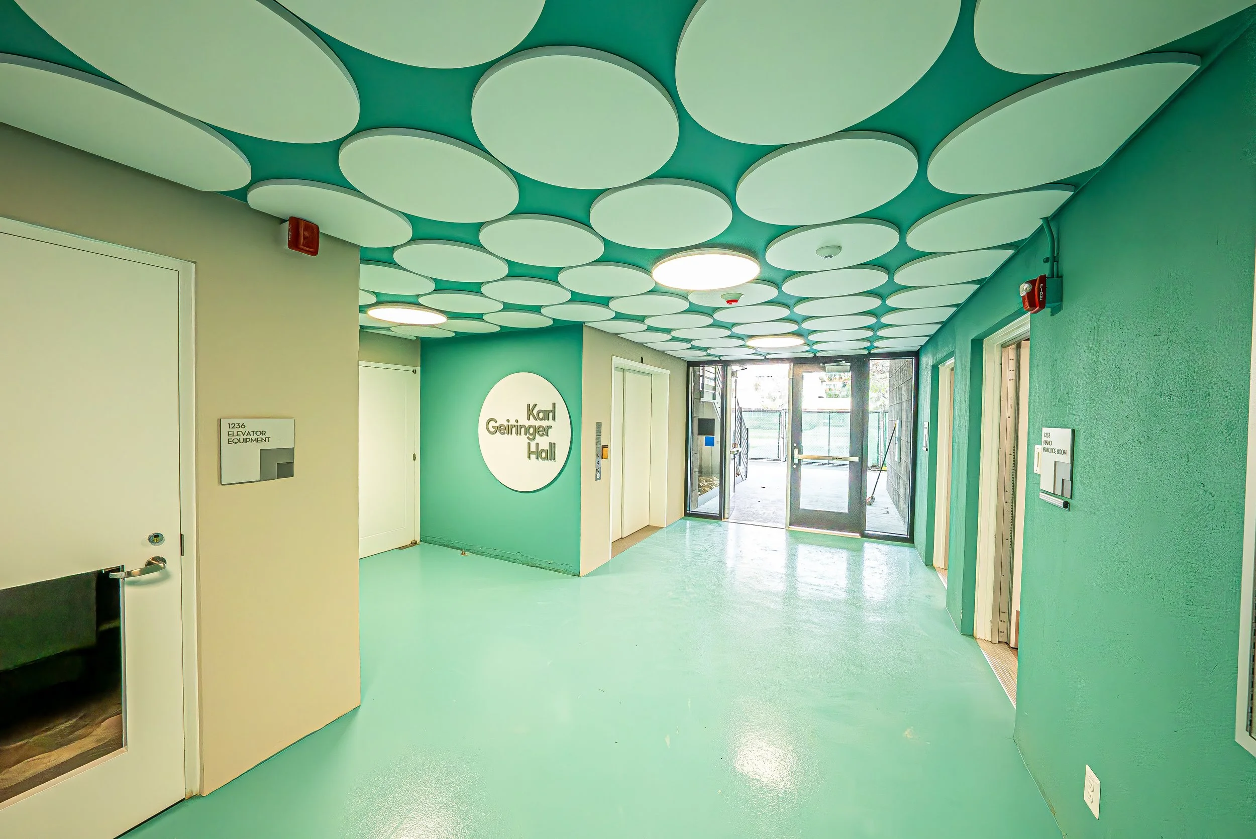 Interior view of the Karl Geiringer Hall with a green floor and ceiling, circular ceiling light fixtures, elevator signage, large glass exit door, and walls painted in shades of green and beige.