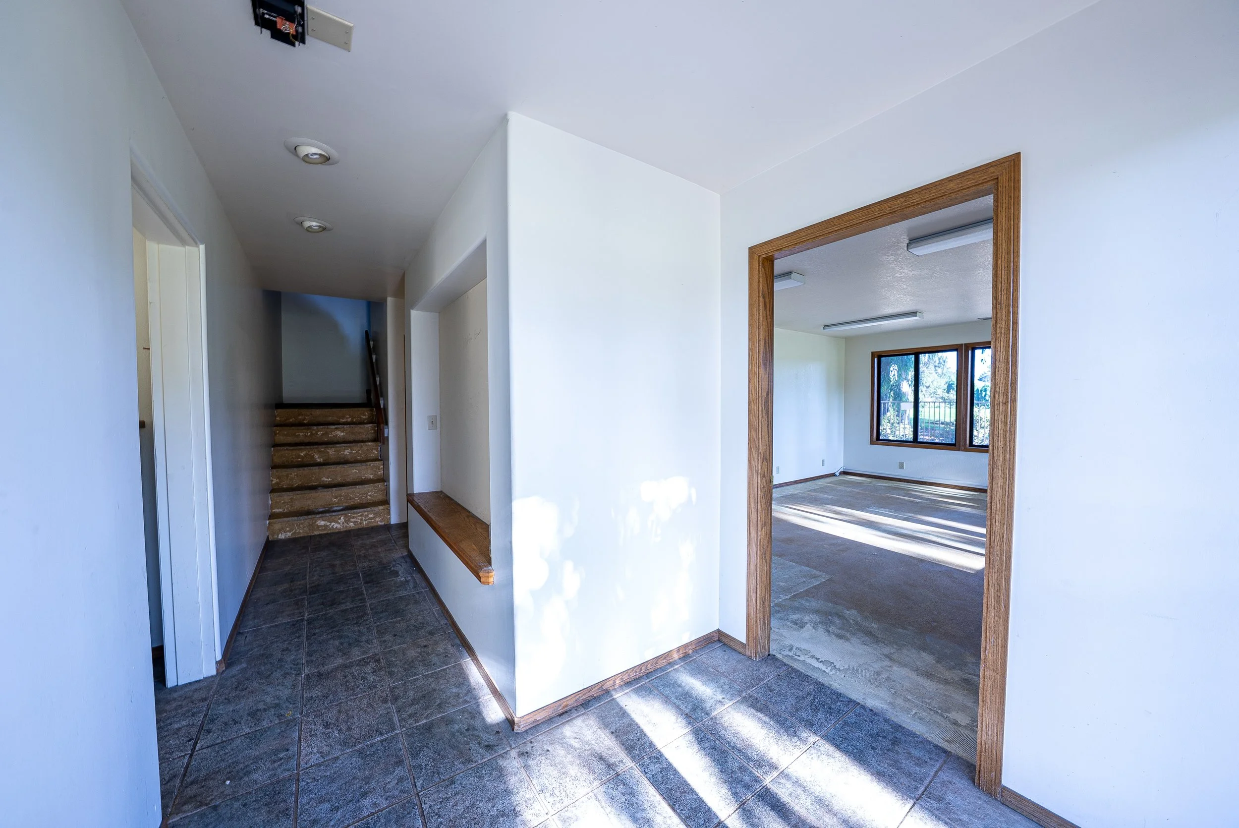Interior of a house with a hallway and an open doorway leading to a room with large windows and sunlight. The hallway has dark tiled flooring, white walls, and a staircase at the end. The doorway trim is wooden.