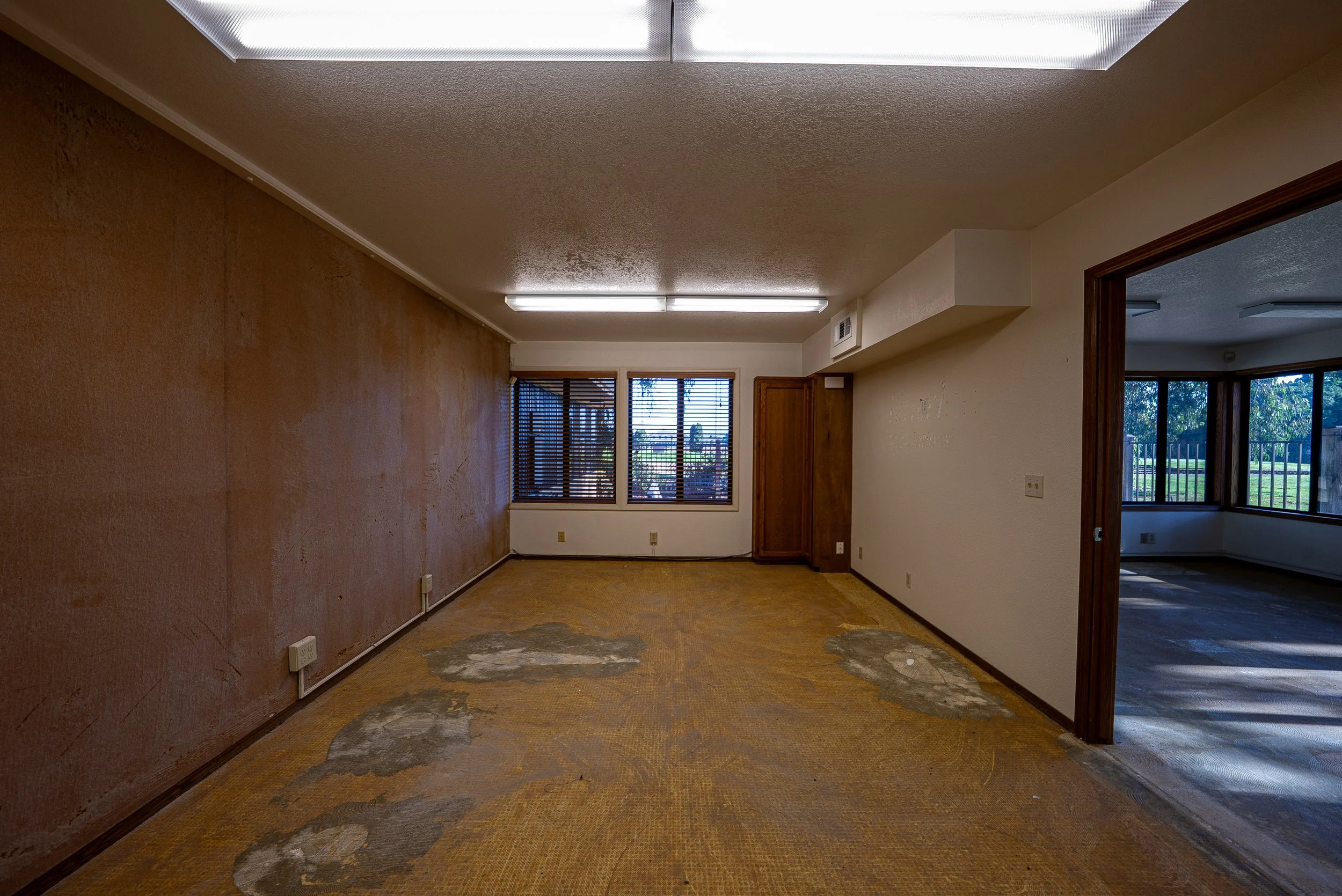 Empty room with brown carpet, textured beige walls, wooden trim, and large windows with blinds, sunlight streaming in.