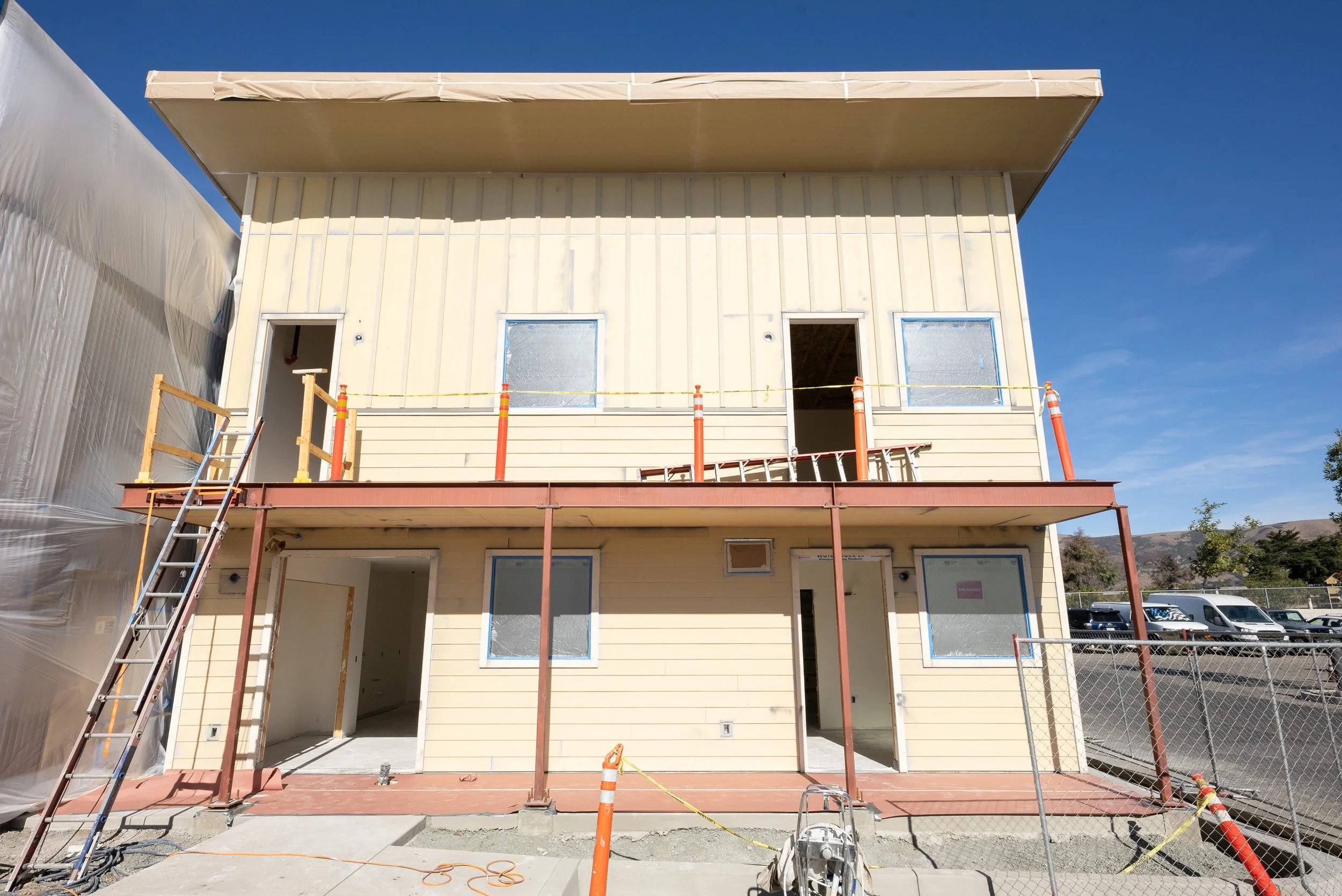 Front view of a two-story house under construction, with beige exterior siding and three open door and window spaces. Construction equipment, ladders, safety cones, and scaffolding are present, with a clear blue sky in the background.