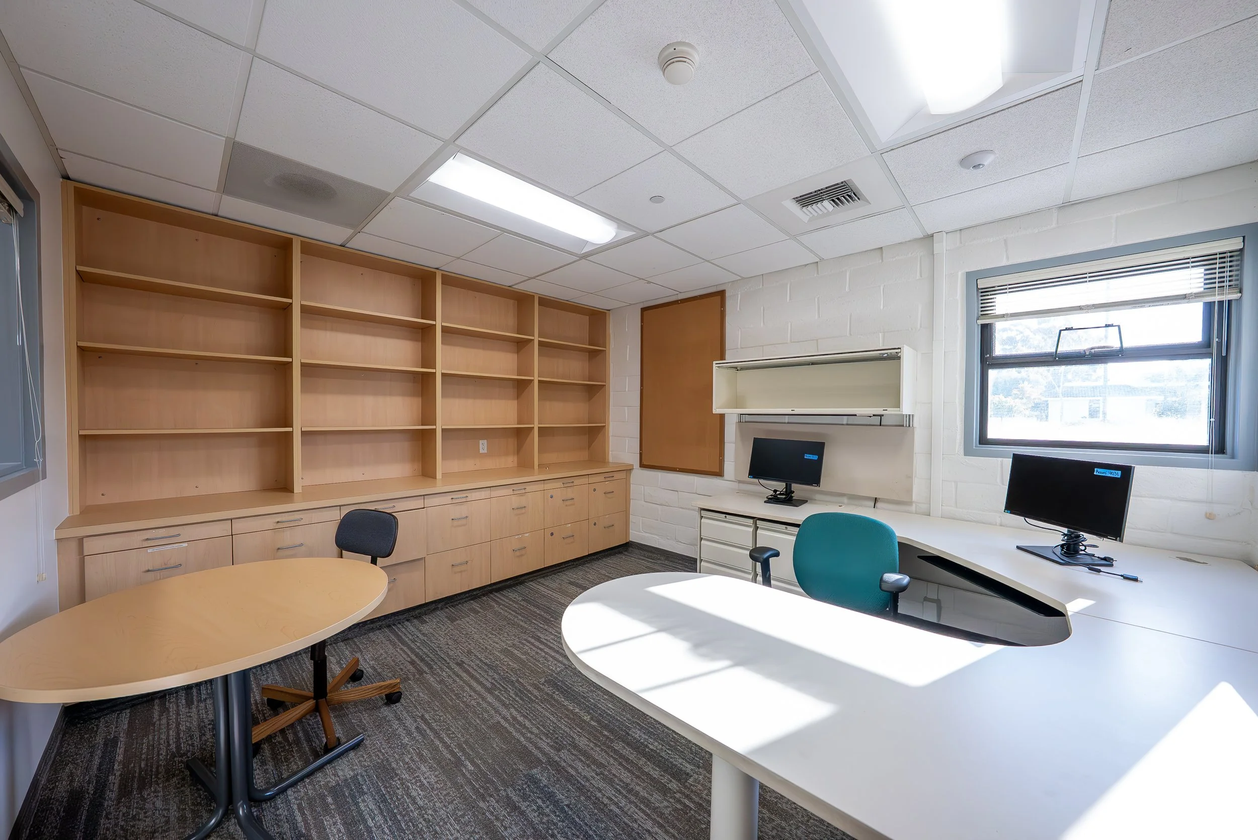 An office room with beige bookshelves, a white brick wall, a large window with blinds, two computer monitors on desks, and various chairs. Bright overhead lighting and carpeted floor.