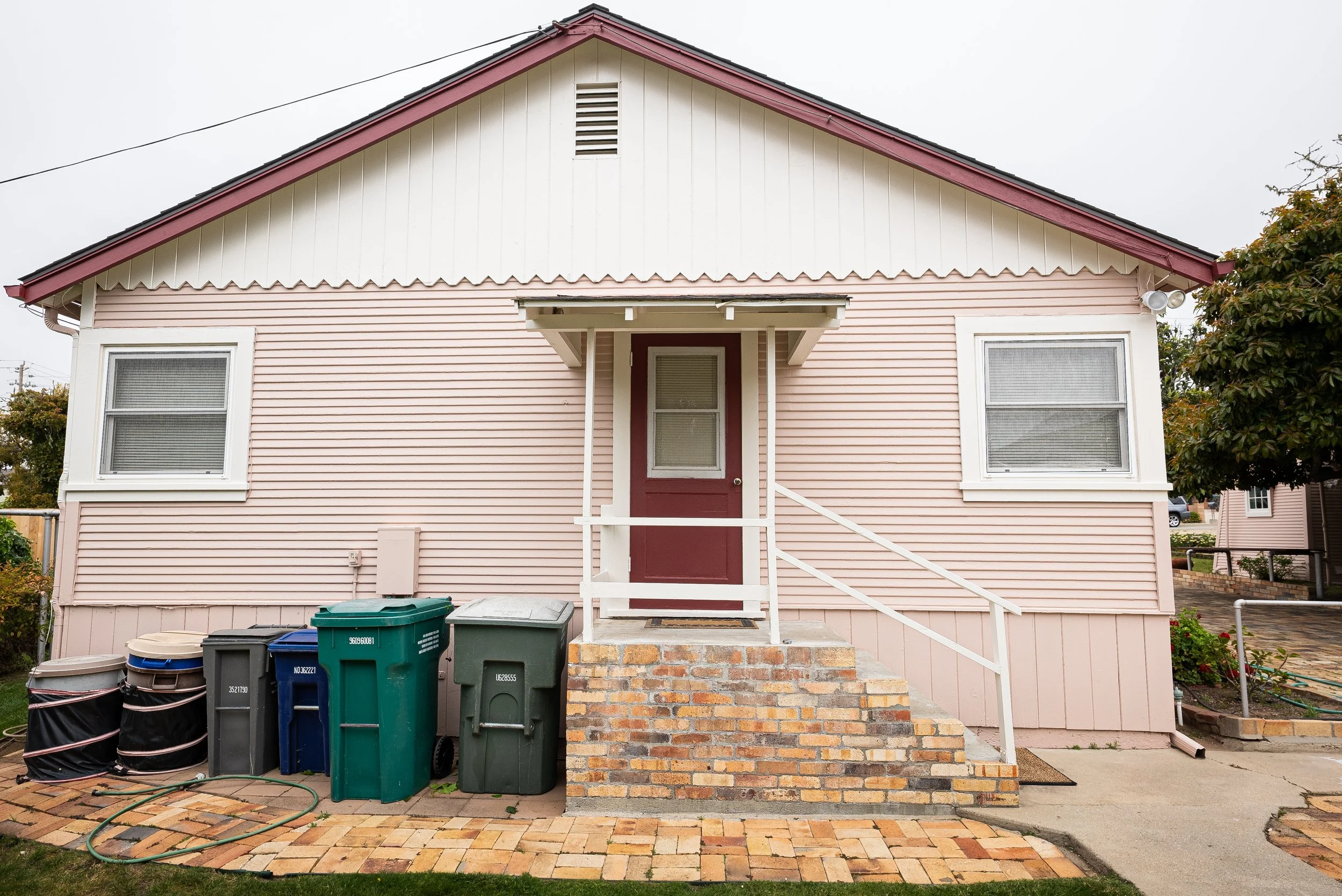Front view of a small pink house with white trim, two windows, a small front porch, and a brick staircase. There are multiple trash and recycling bins in the front yard, and a cloudy sky overhead.