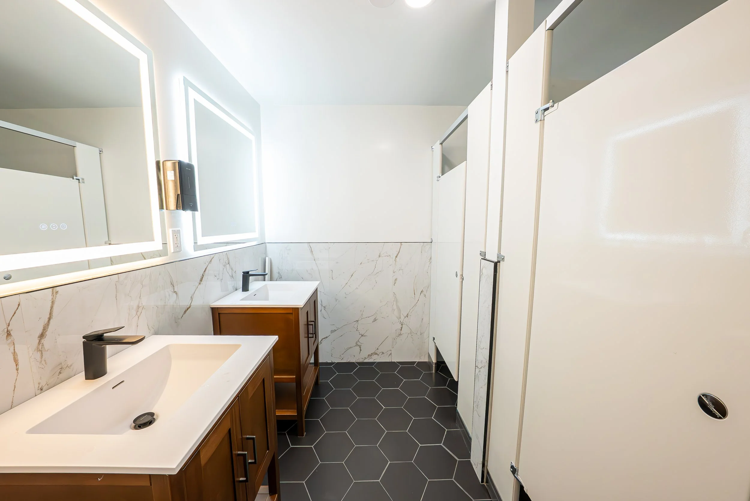 A modern bathroom with white walls, marble backsplash, black hexagon tile floor, wooden vanity with a white sink, and a large backlit mirror.