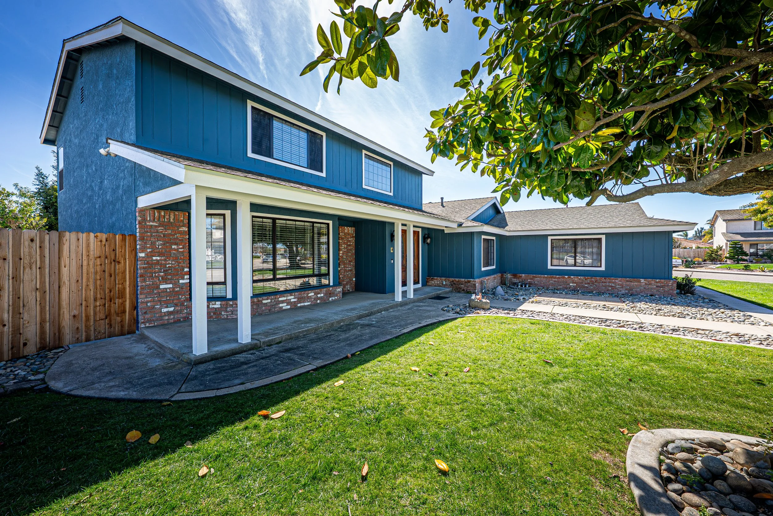 View of a two-story house with blue siding, white trim, and brick accents, with a yard featuring green grass, a tree, and a small rock garden under a clear blue sky.