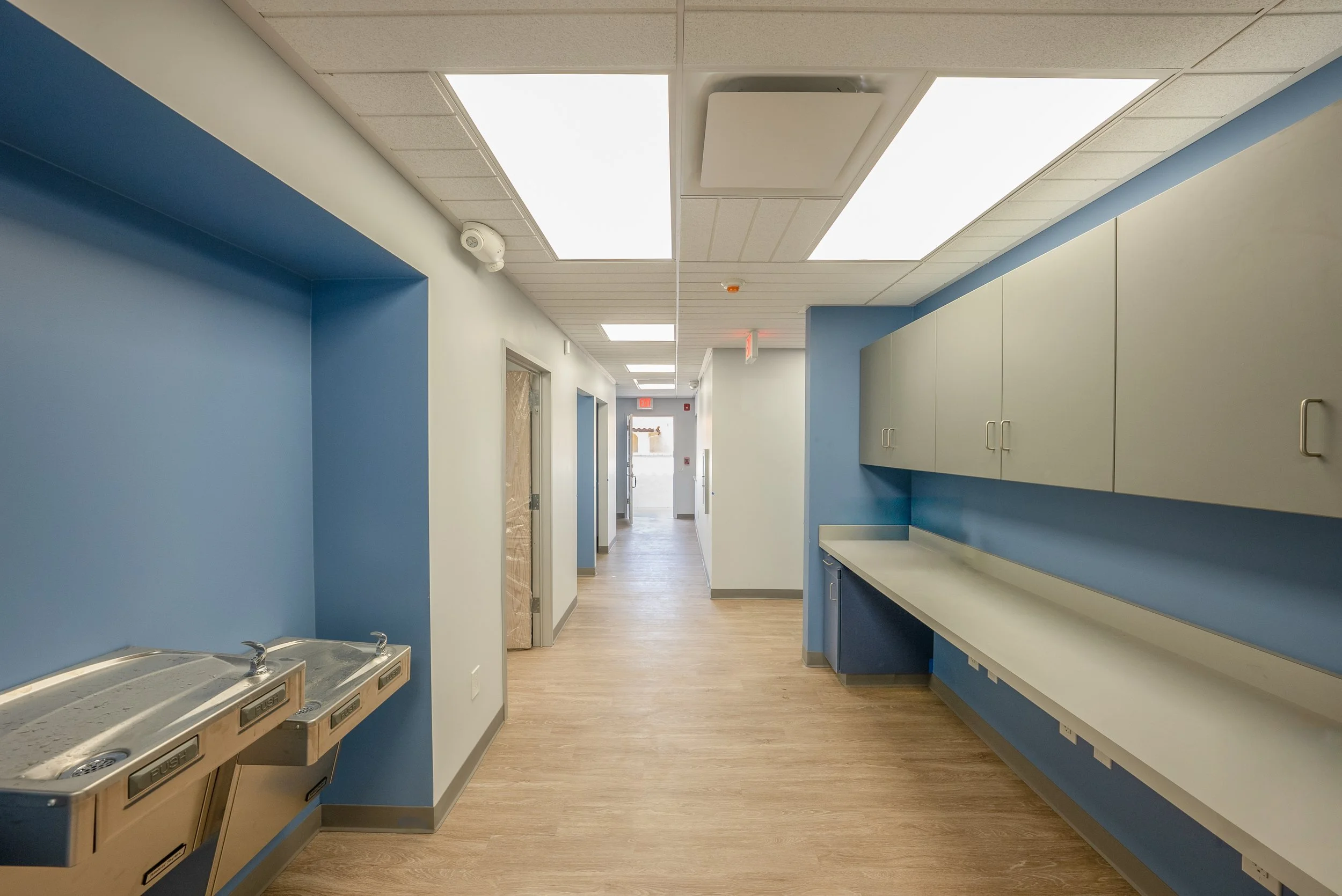 Hospital hallway with blue and white walls, ceiling lights, water fountain, cabinets, and an exit door in the distance.