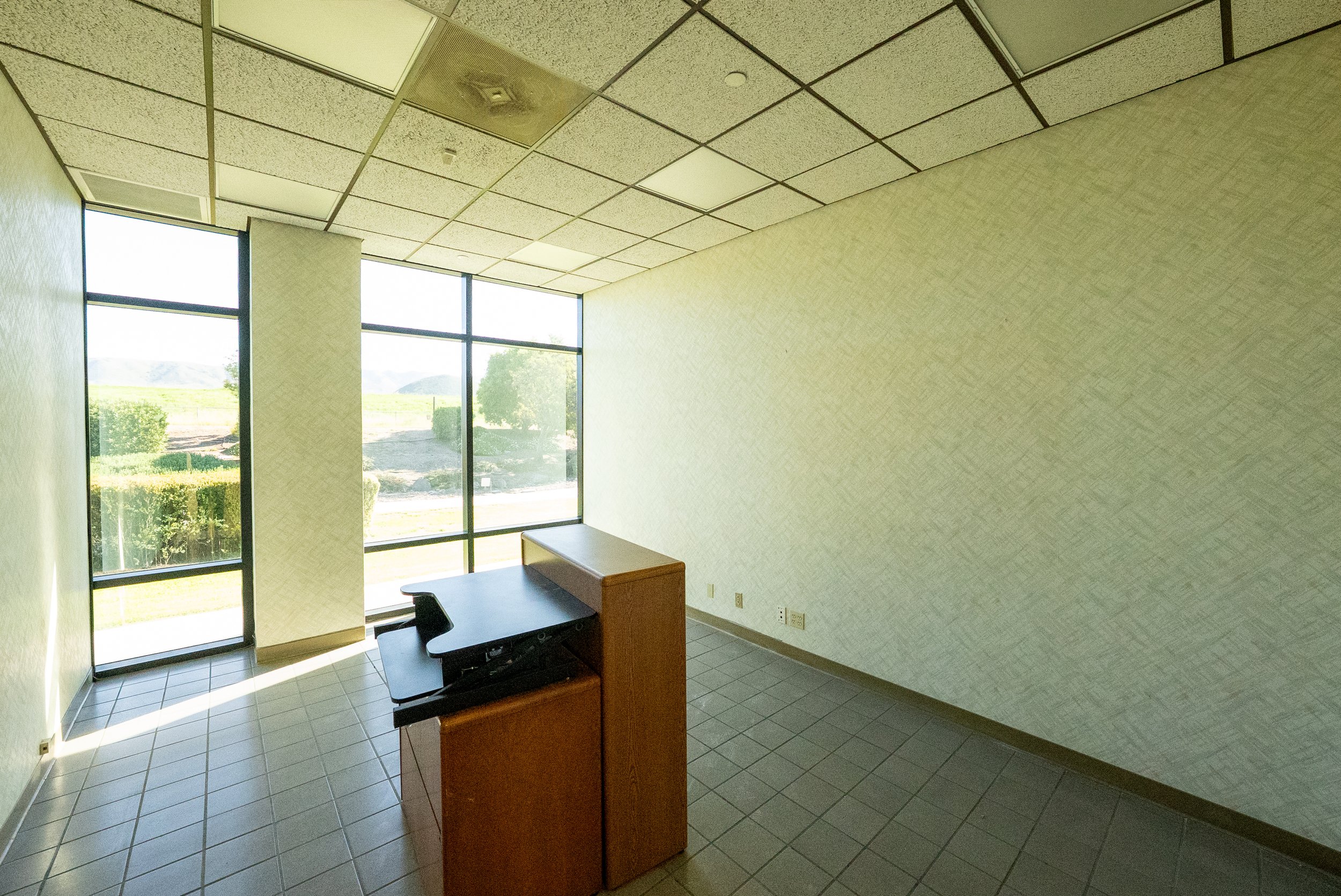 An interior view of an empty office or waiting room with beige textured walls, a tiled floor, and large windows letting in natural light. There is a desk, a chair, and a small wooden cabinet.