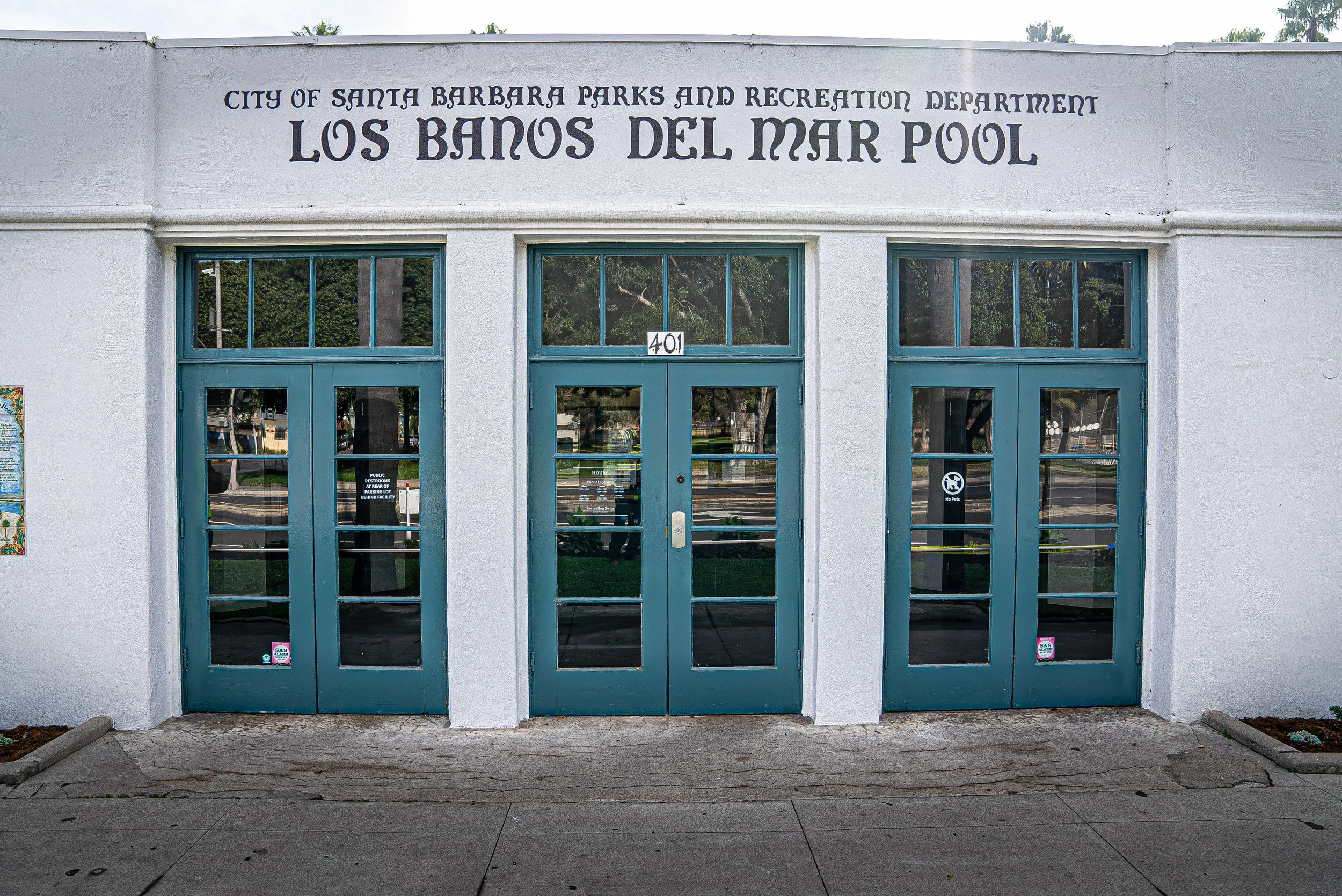 The entrance of the Los Banos del Mar Pool building in Santa Barbara, California, with teal double doors and a sign with the address number 401 above the doors, under a white wall with black lettering.
