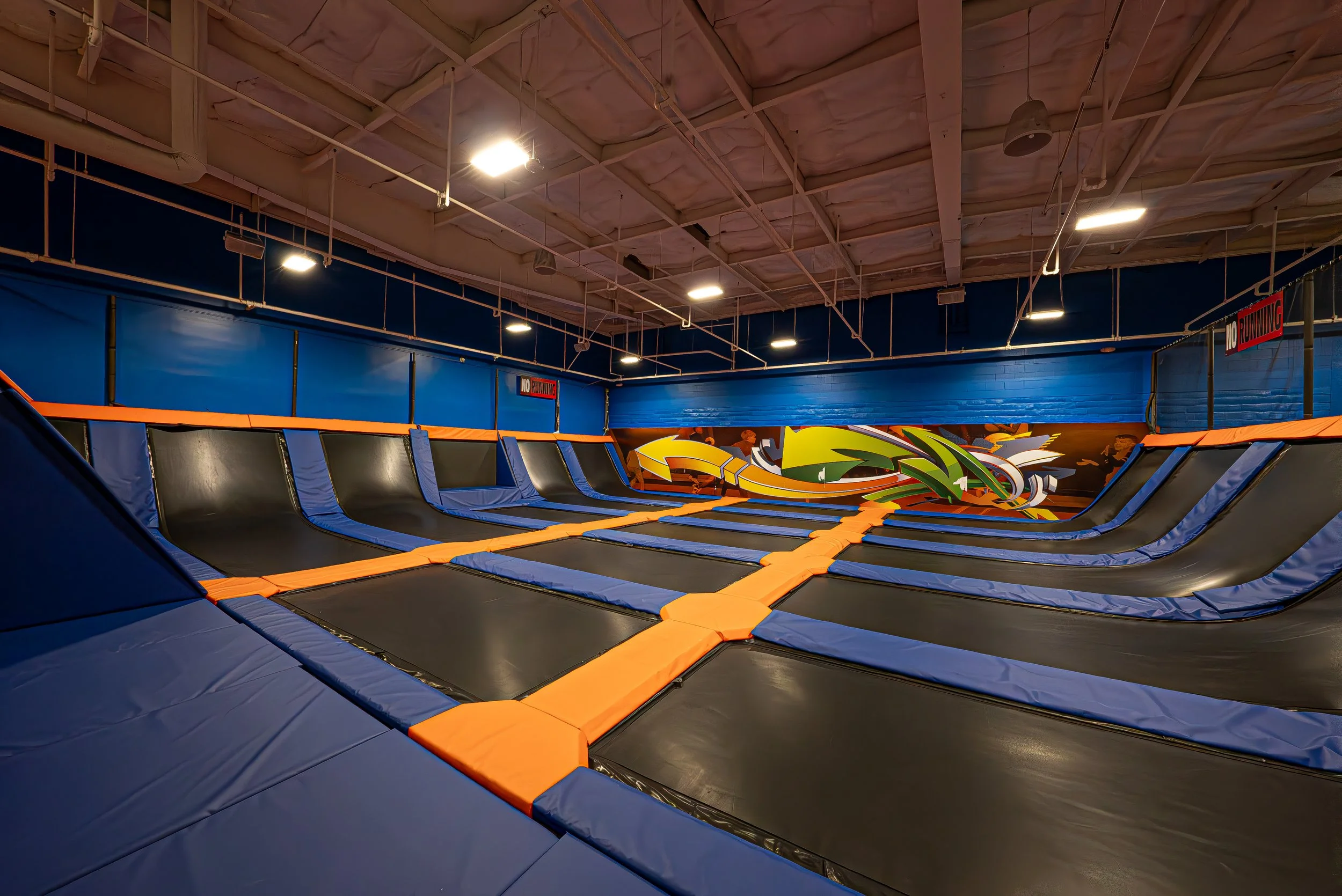 Indoor trampoline park with multiple interconnected trampolines bordered by blue and black padding, colorful graffiti mural on the far wall, and a sign that reads 'No Running' on the blue wall.