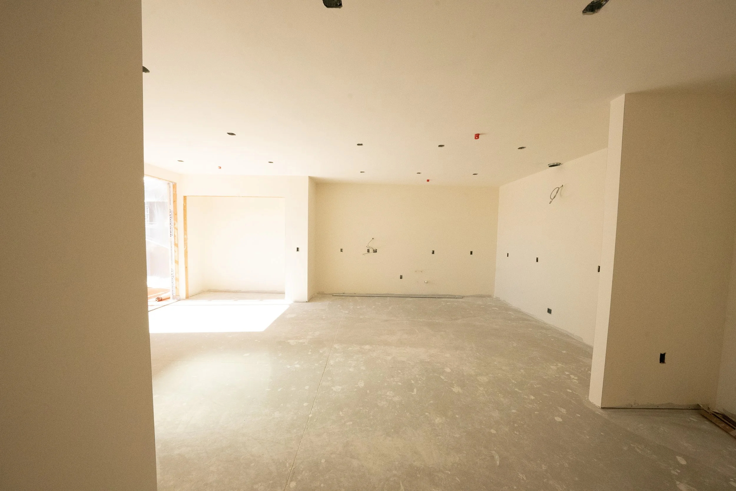 An empty room under construction with white walls, unfinished flooring, and visible electrical outlets and wiring. There is a large window letting in natural light.