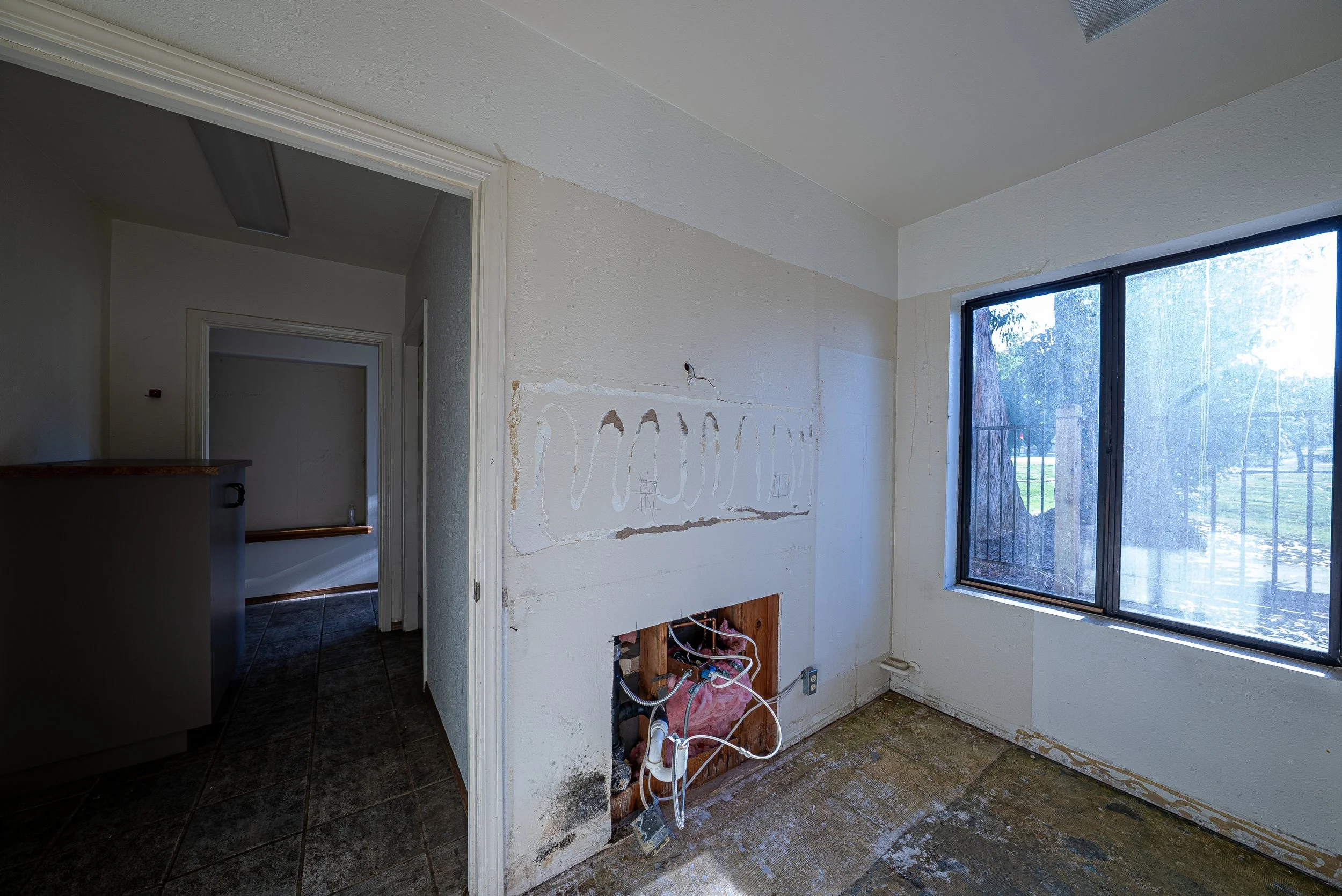 Interior room undergoing renovation with exposed plumbing, no cabinet or appliances, and a window showing an outdoor scene.