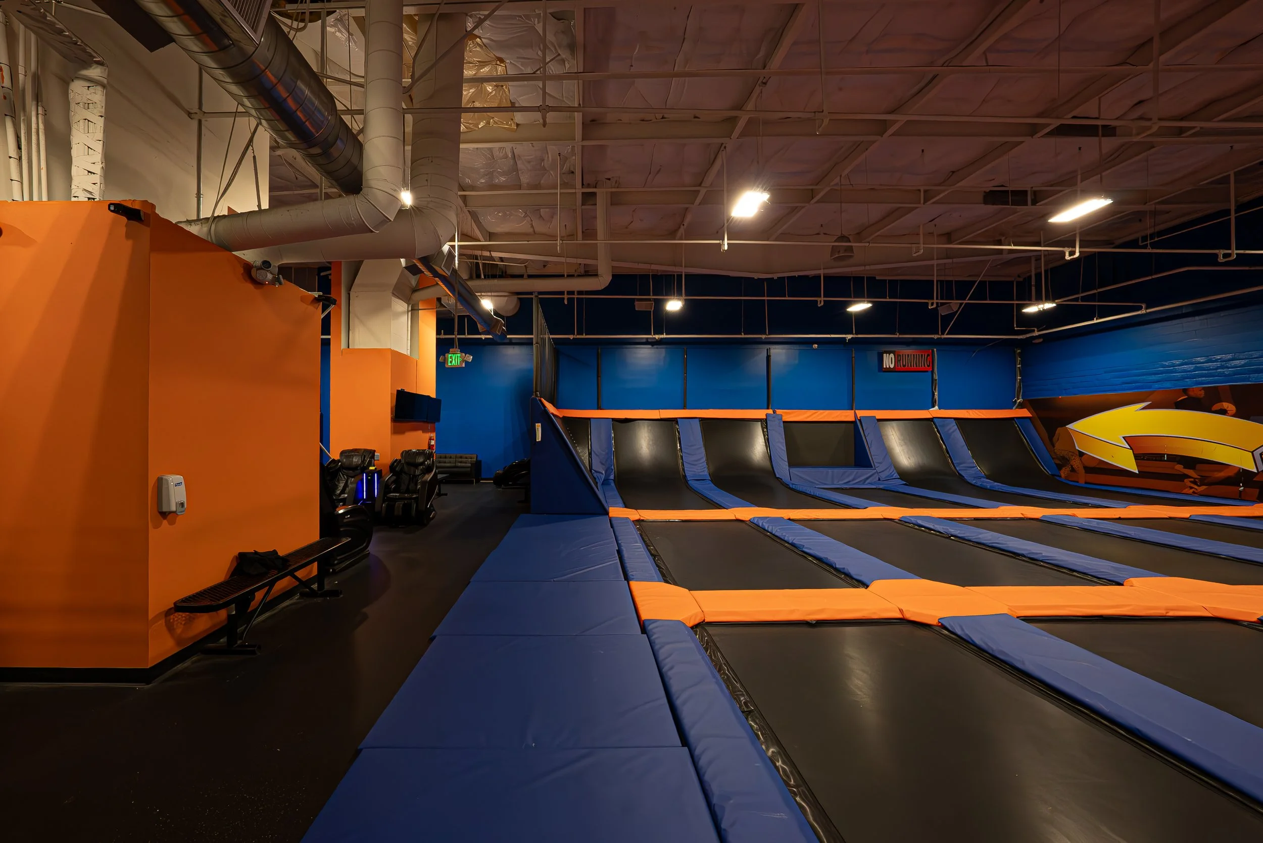 Indoor trampoline park with blue and orange padded trampolines, foam pits, and wall pads, under a ceiling with exposed ductwork and lighting.