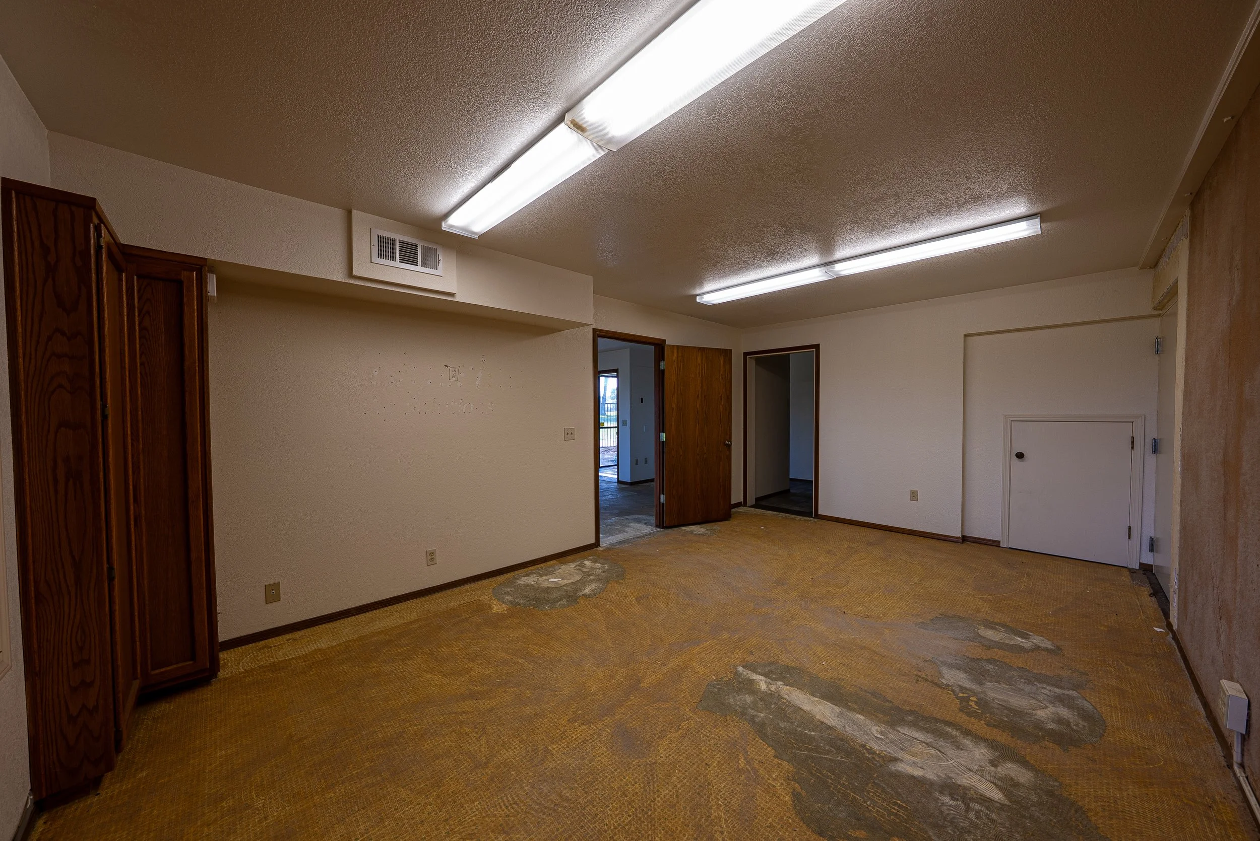 Empty room with a damaged carpet, open wooden doors, and ceiling fluorescent lights.