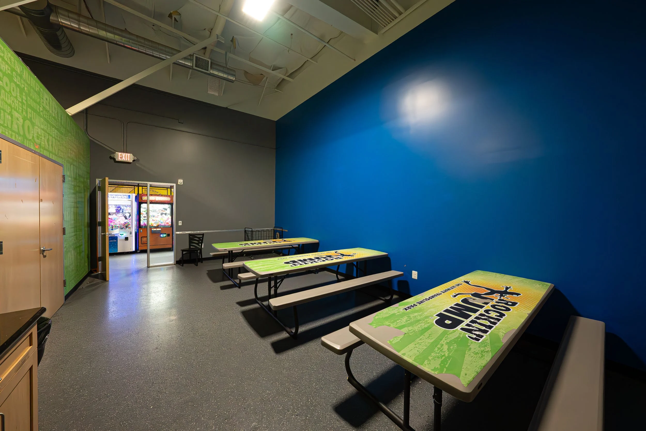 Indoor recreational area with blue wall, partitioned tables with advertising to promote a game or event, vending machines in the background, combination of gray and green walls, and ceiling with exposed ducts and lighting.