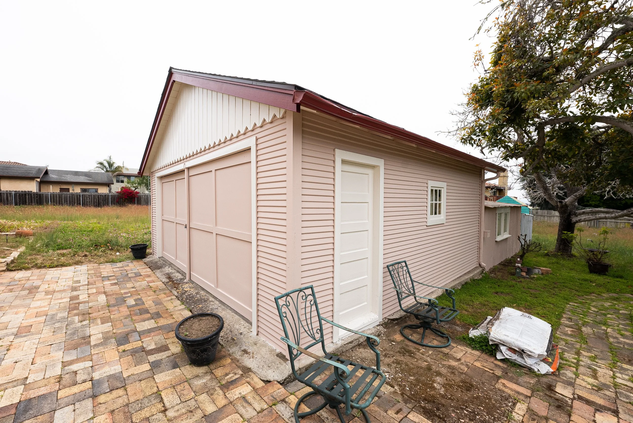 Backyard shed with pink siding, white door, and small window; brick patio and garden chairs in the foreground; tree and neighboring houses in the background.