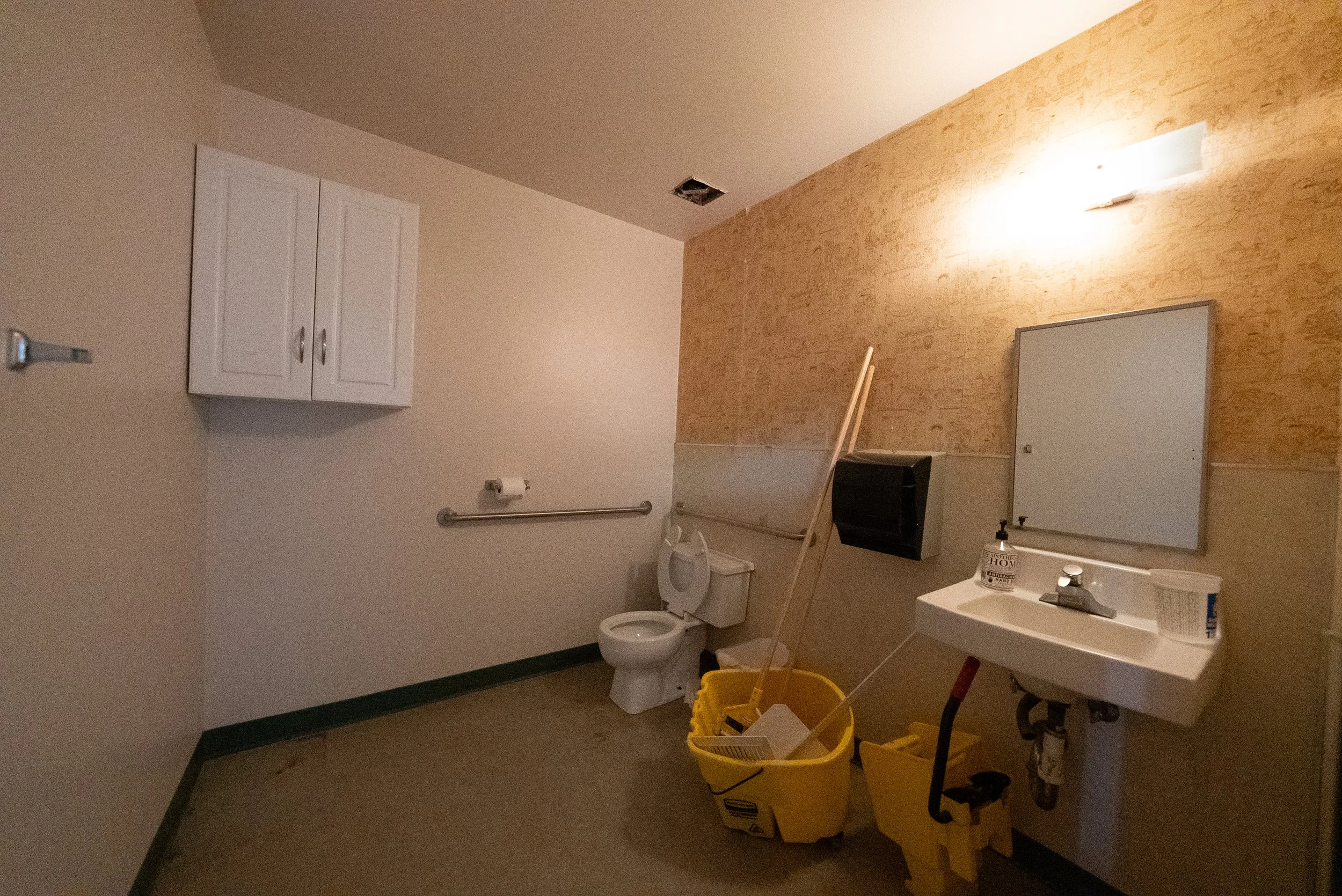 An indoor medical or clinic room with beige and white walls, a mirror above a sink, a toilet, a white cabinet, and cleaning supplies including mop buckets and mop sticks.
