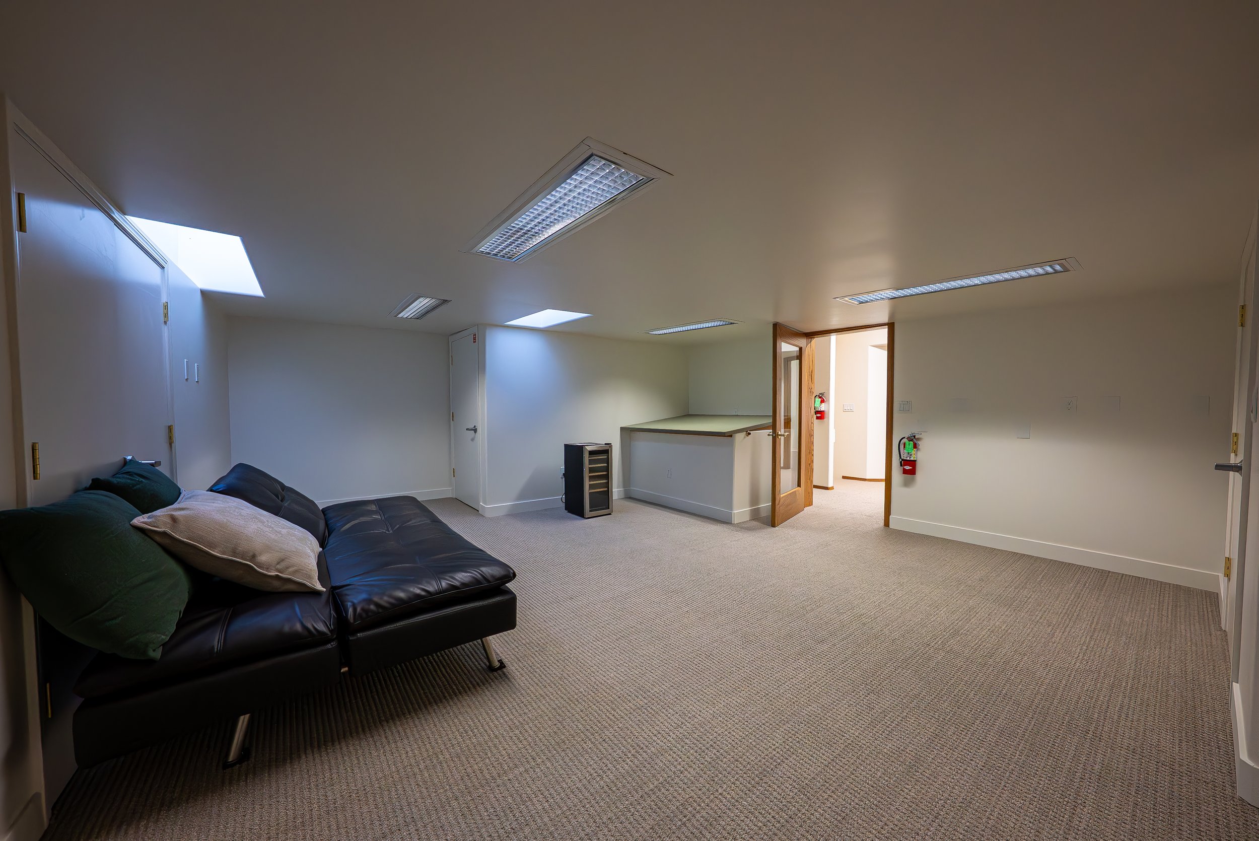 An interior room with a black leather sofa, pillows, fluorescent ceiling lights, small windows, and an open door leading to a hallway with fire safety equipment.